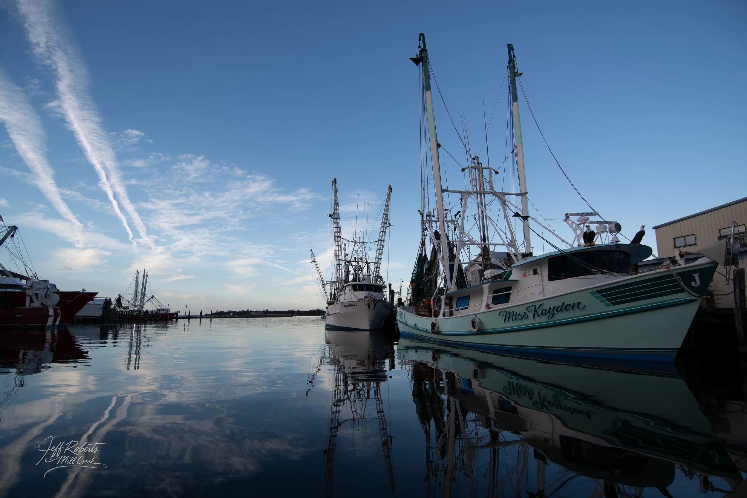 Boats docked at a marina with calm water reflecting the vessels and a blue sky with wispy clouds.
