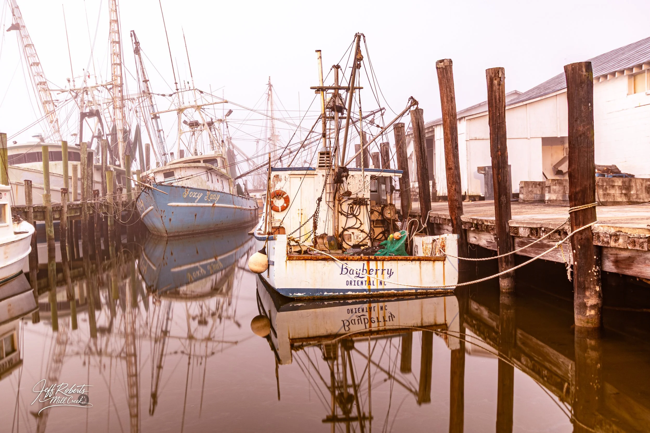 Several boats docked at a pier with calm water reflecting their images. The boats vary in size and condition, with some appearing old and worn. Wooden pilings and a white building are also visible in the background.