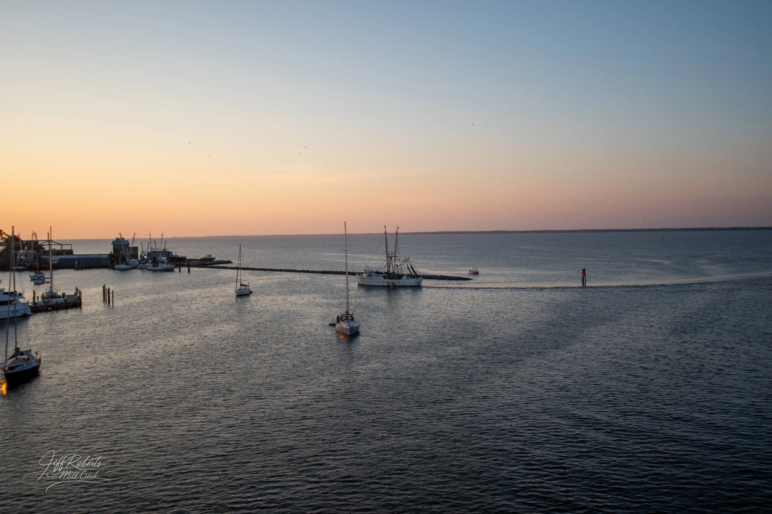 A harbor at sunset with sailboats docked and a boat leaving the pier, calm water, and a clear sky with a gentle gradient from pale near the horizon to darker blue at the top.