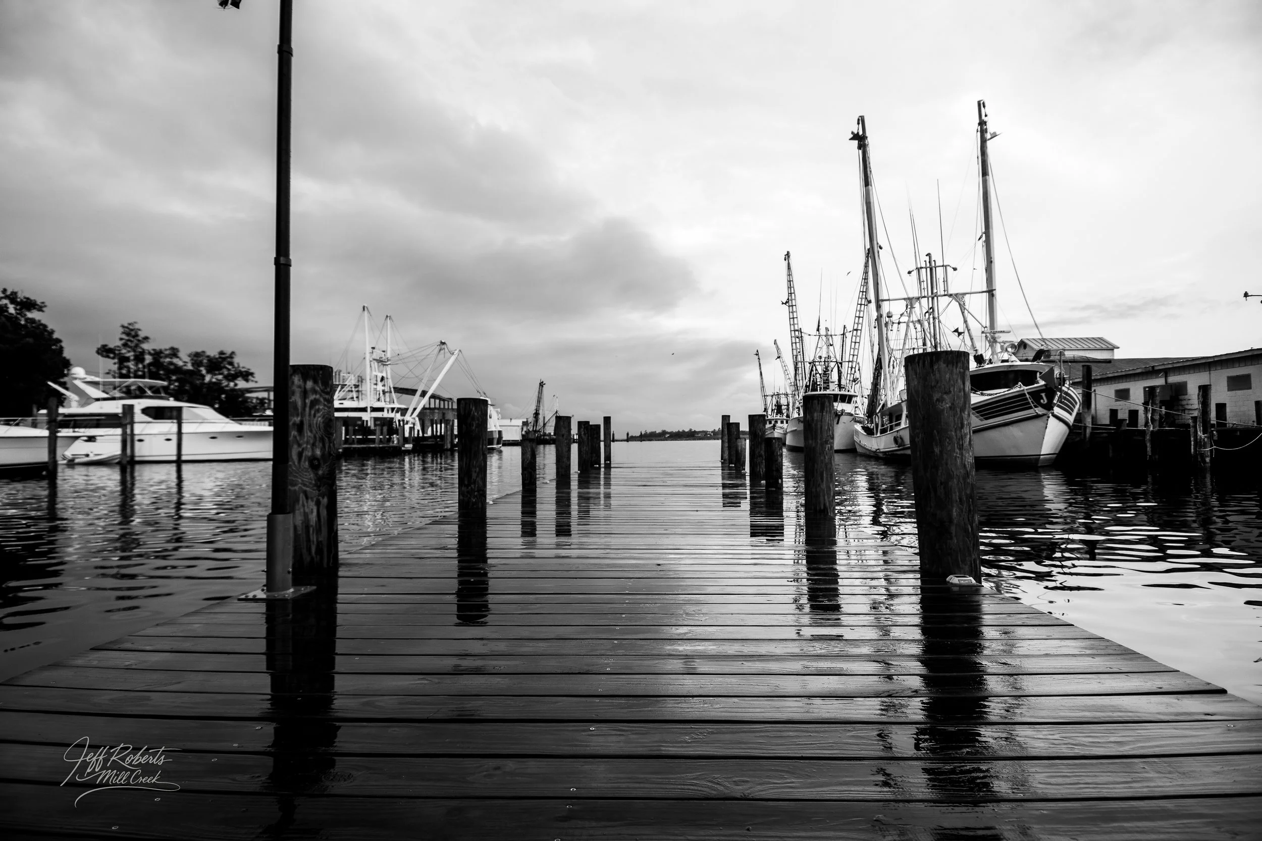 Black and white photo of a dock with boats and yachts moored on both sides, with clouds overhead and reflections on the water.