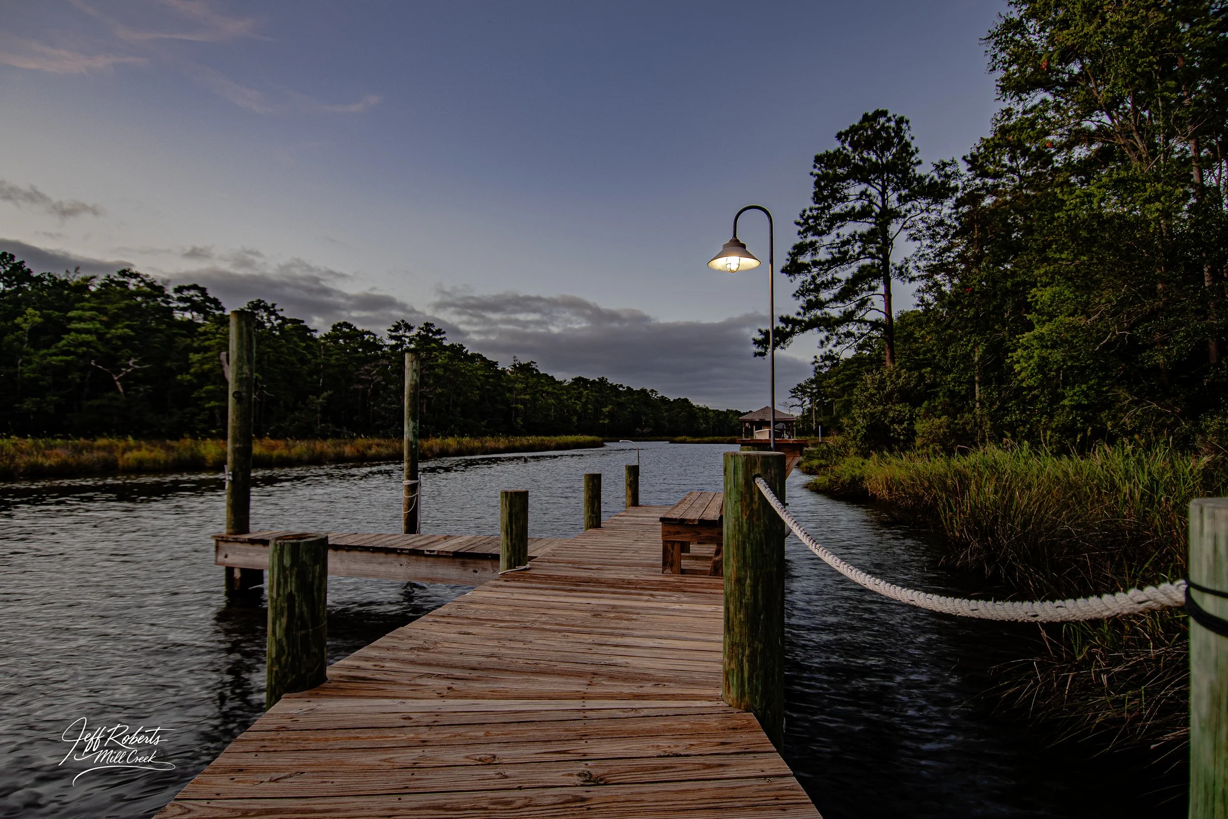 A wooden dock extending over a body of water at dusk, with a lamppost lit along the dock, surrounded by trees and vegetation.