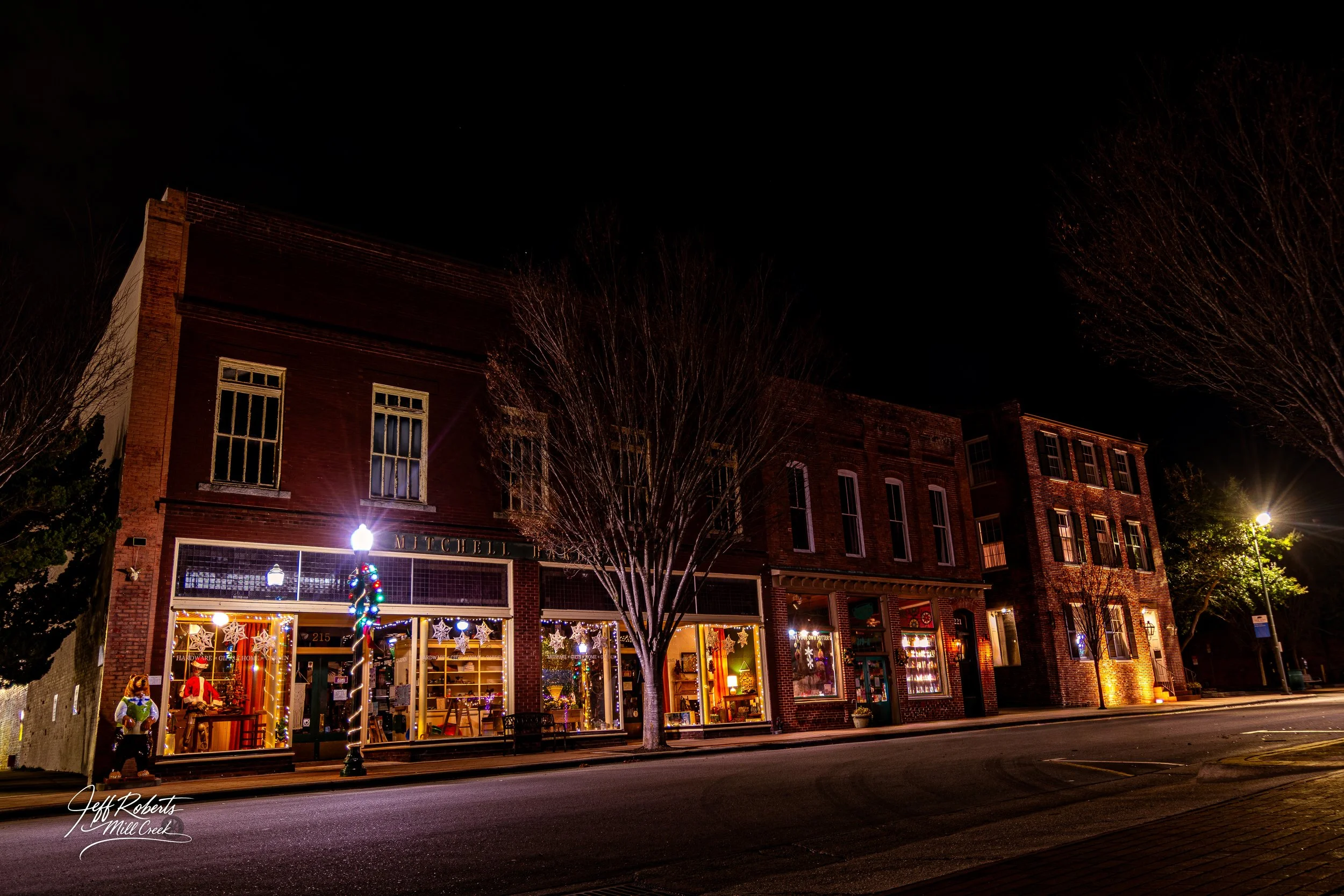 Night view of a downtown street decorated with holiday lights and ornaments, featuring brick buildings, leafless trees, and street lamps illuminating the scene.