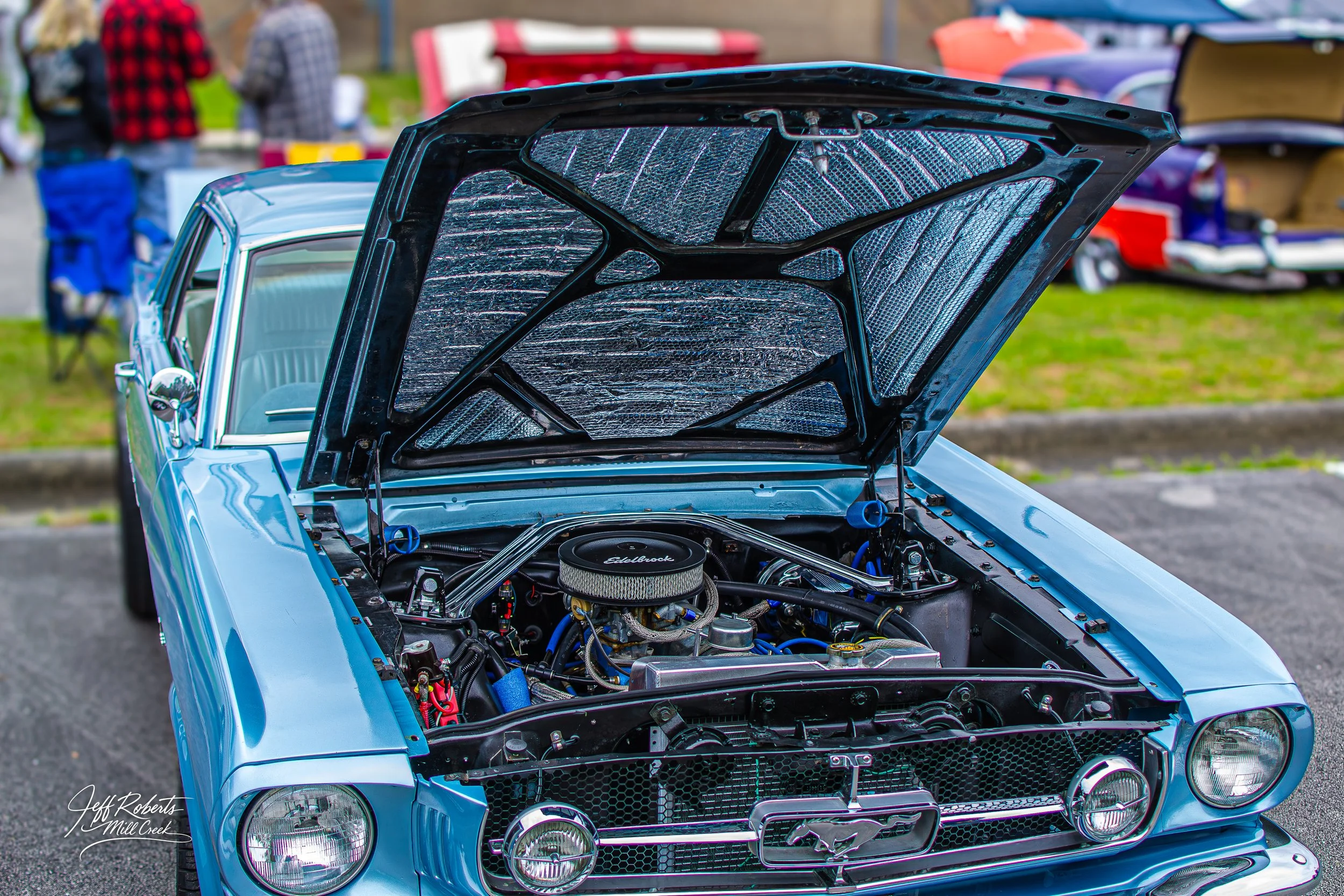 A blue classic Ford Mustang with its hood open, showcasing the engine at an outdoor car show.