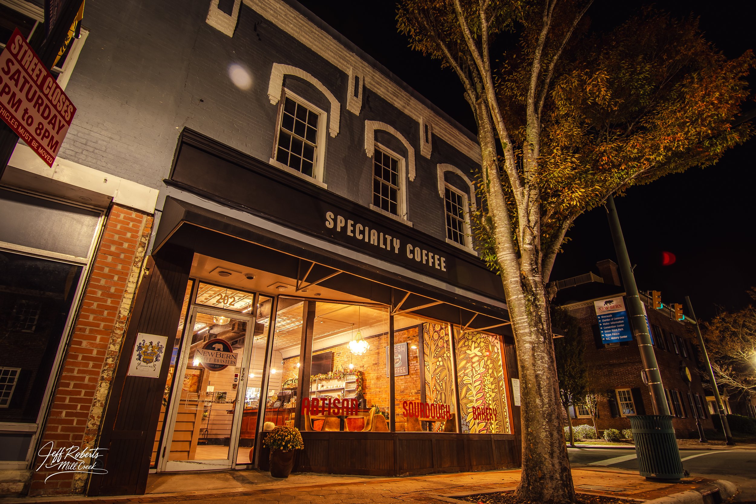 A nighttime street view of a coffee shop named 'Specialty Coffee' with warm interior lighting, large glass windows displaying signs like "Artisanal," "Sourdough," and "Bakery," a potted plant outside, and a tree partially covering the building. Stree