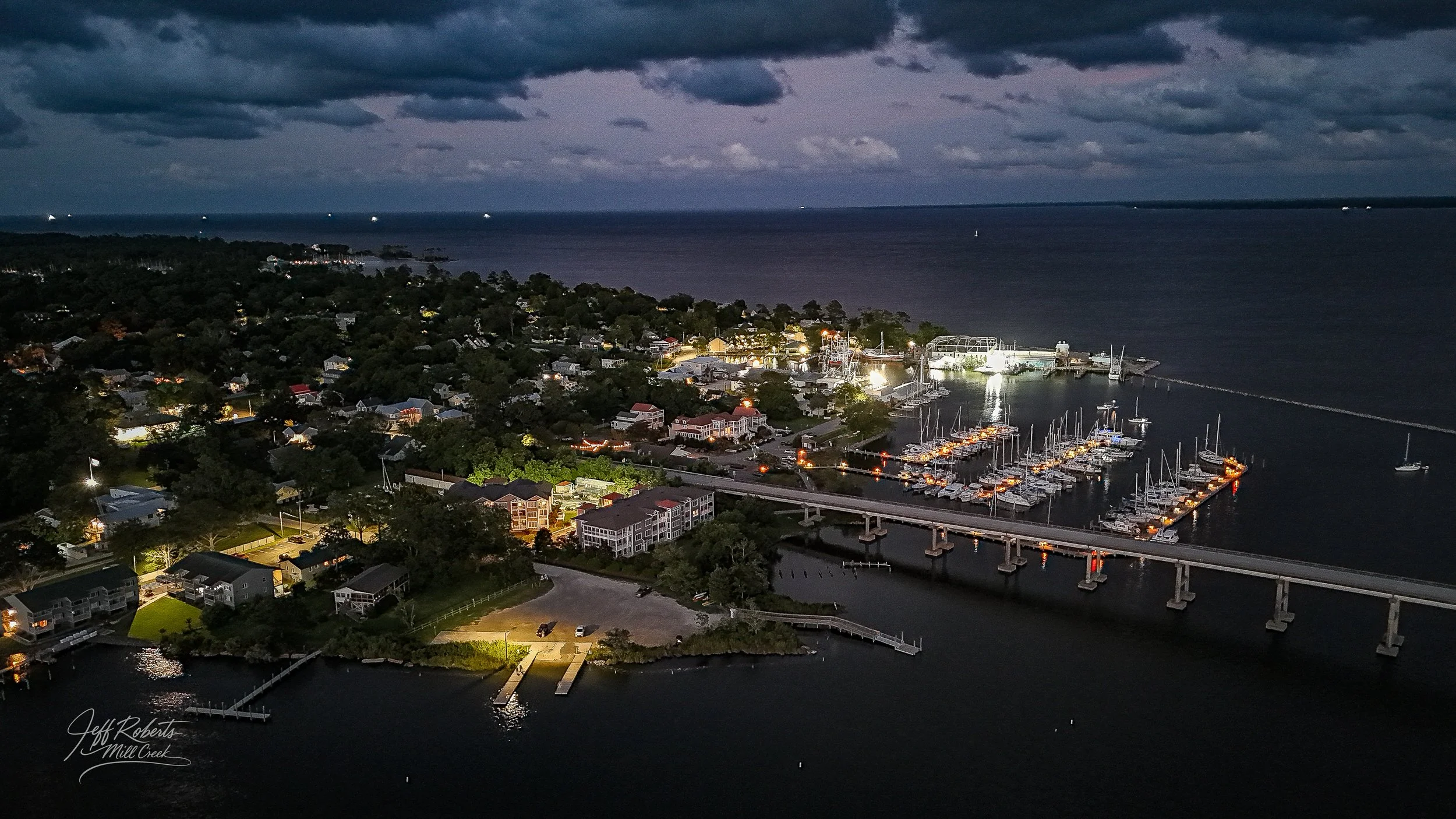 Aerial night view of a coastal town with illuminated houses, marina filled with boats, a bridge, and a dark ocean under cloudy sky.