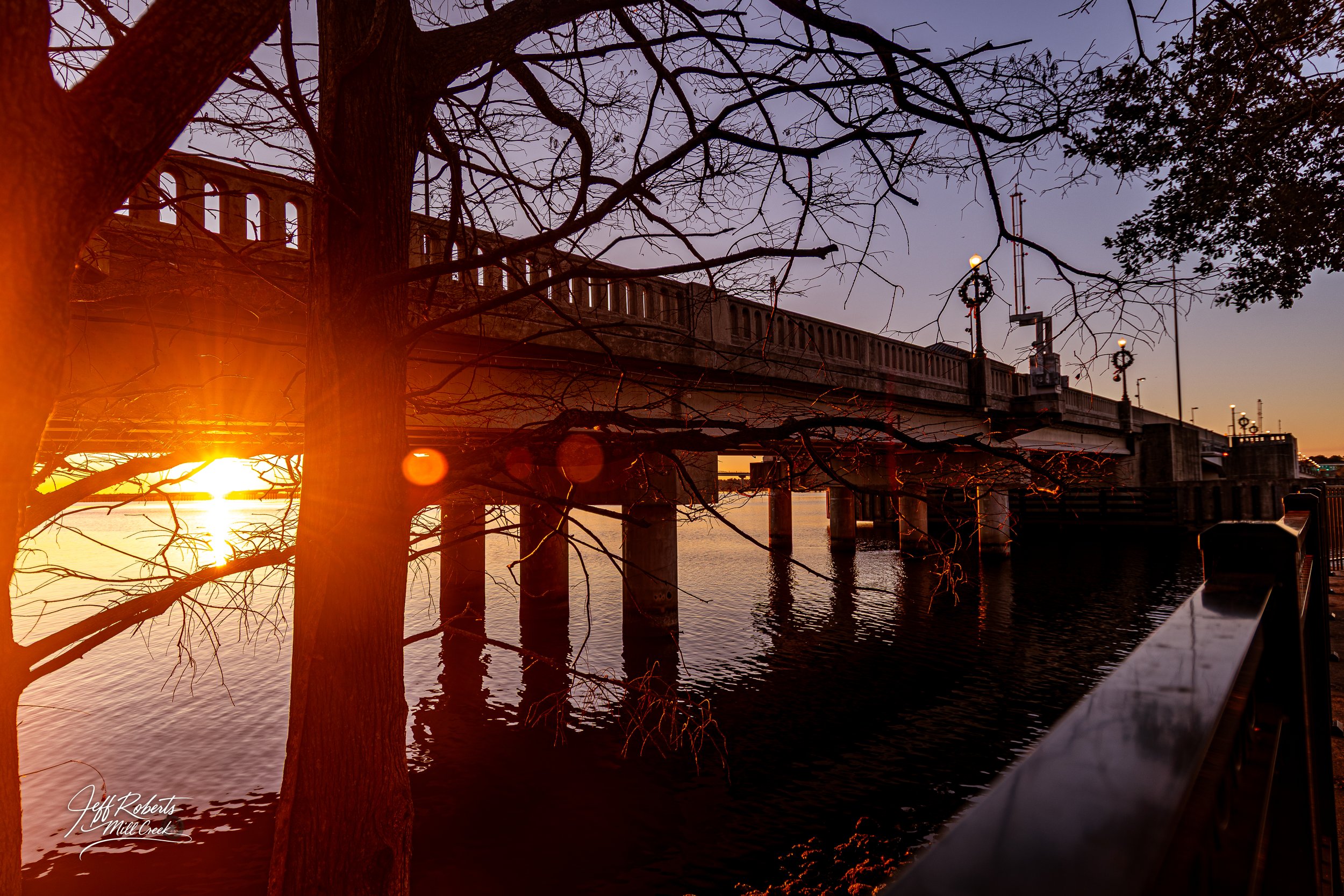 A bridge over a body of water during sunset, with leafless trees in the foreground and ornate street lamps on the bridge, creating a peaceful scene.