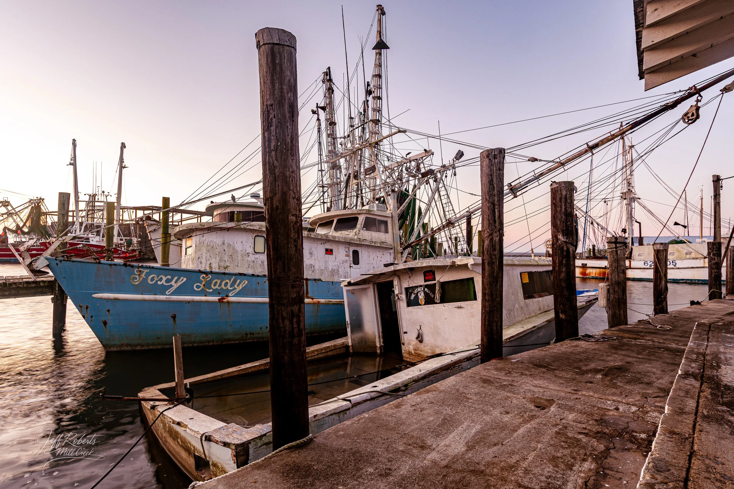 A dock with various old and abandoned boats, including a blue boat named 'Foxy Lady,' floating on calm water with a background of a soft pink and purple sunset sky.