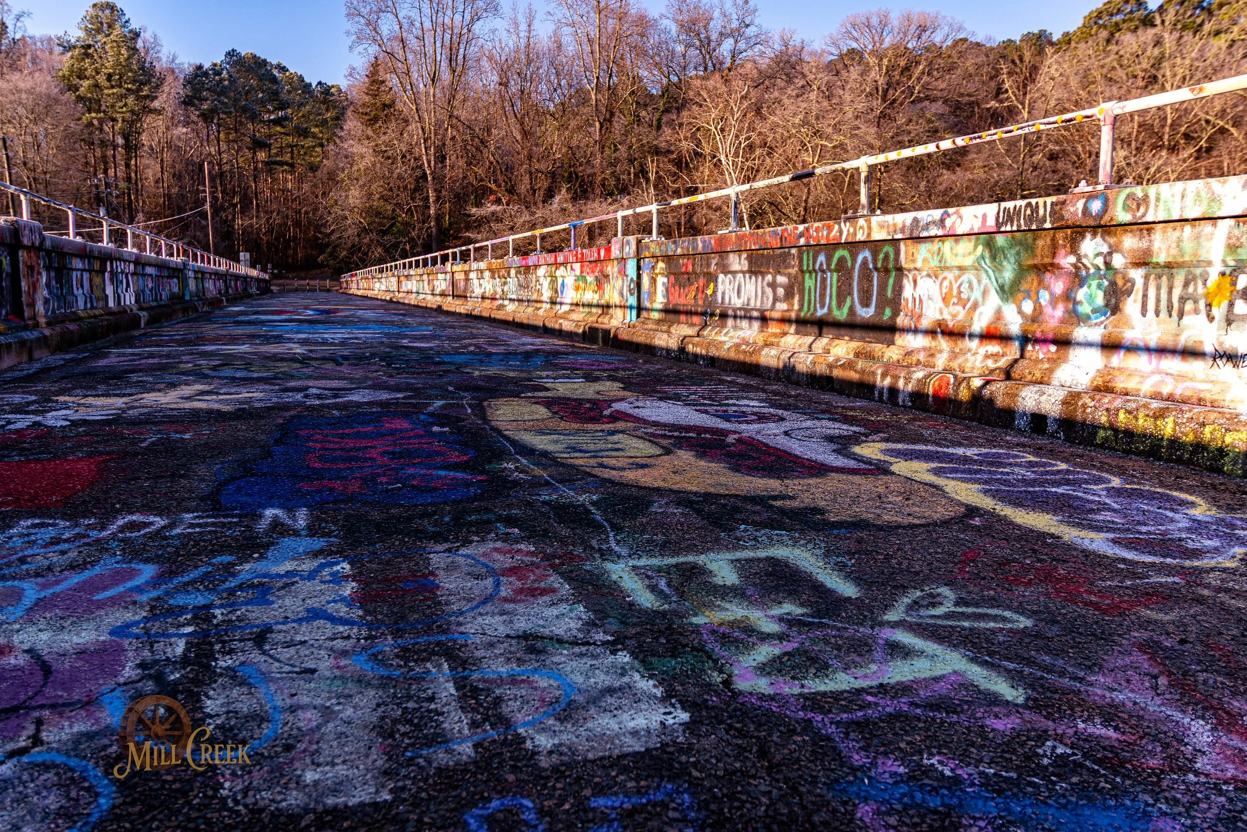 A graffiti-covered bridge with colorful chalk drawings on the pavement and a wooded area in the background.