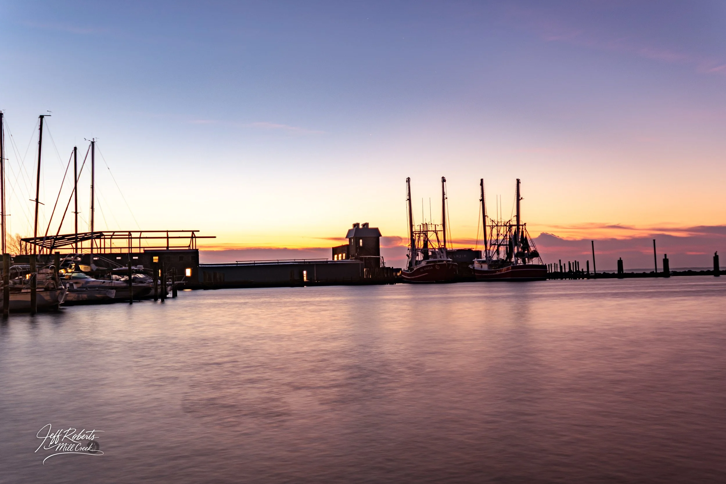 Calm harbor at sunset with boats docked, a small building on the pier, and a colorful sky