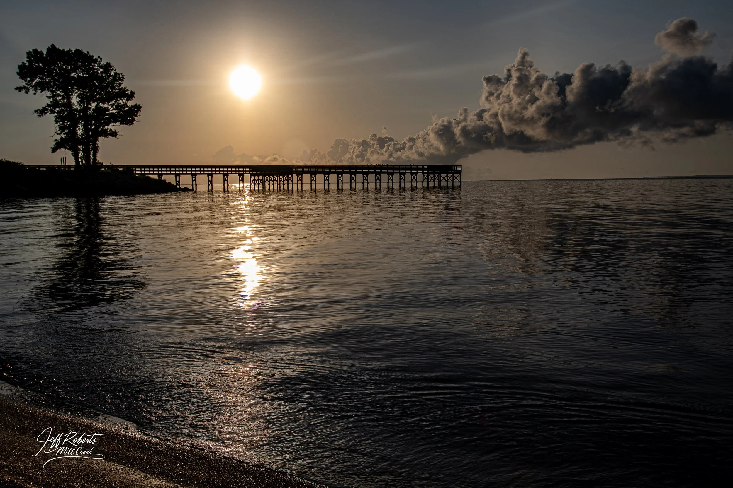 Sunset over a body of water with a wooden pier extending into the distance, a large tree on the left, and dark clouds in the sky.
