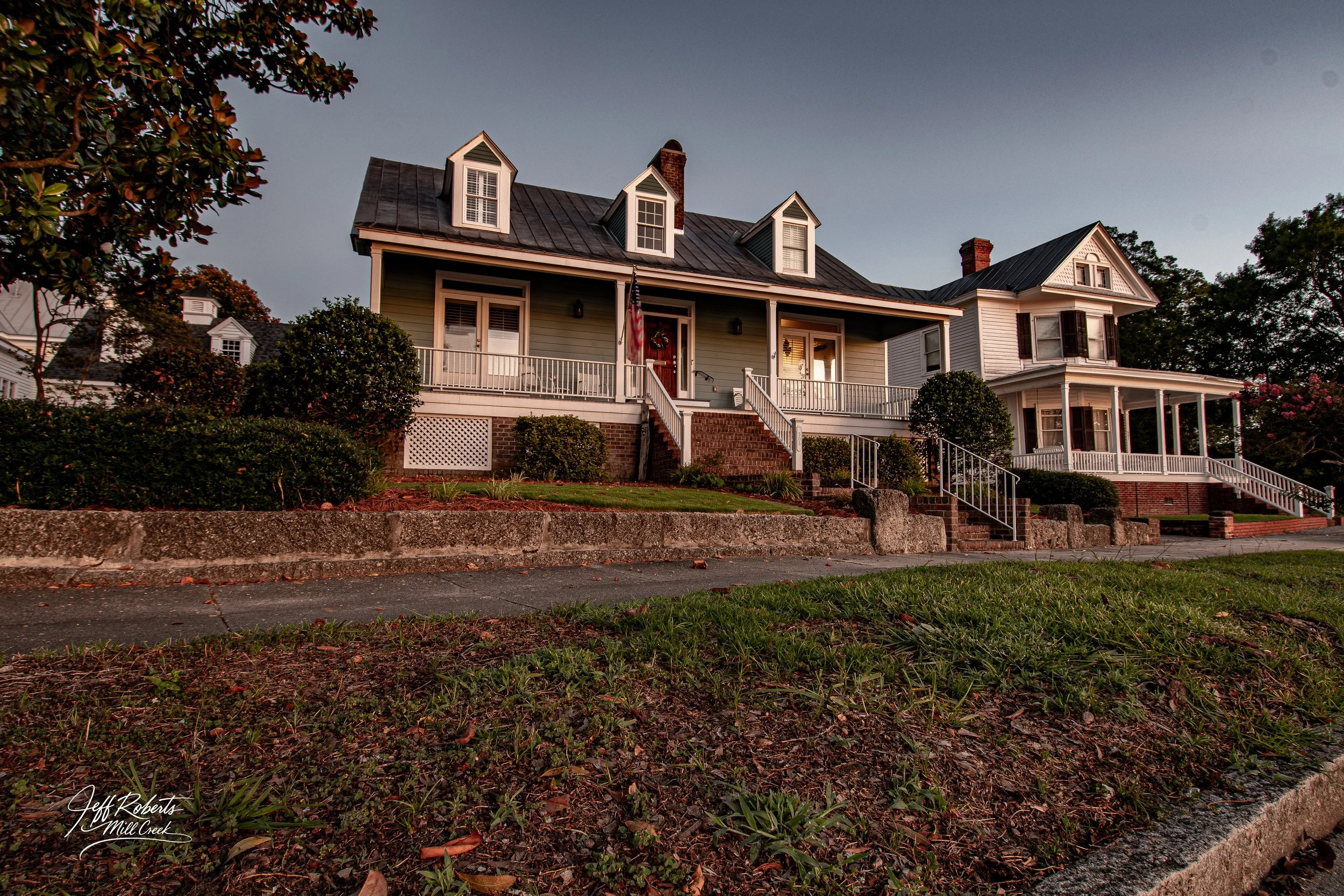 A multi-story house with a front porch, trim and shutter details, surrounded by a well-kept yard and trees during sunset.