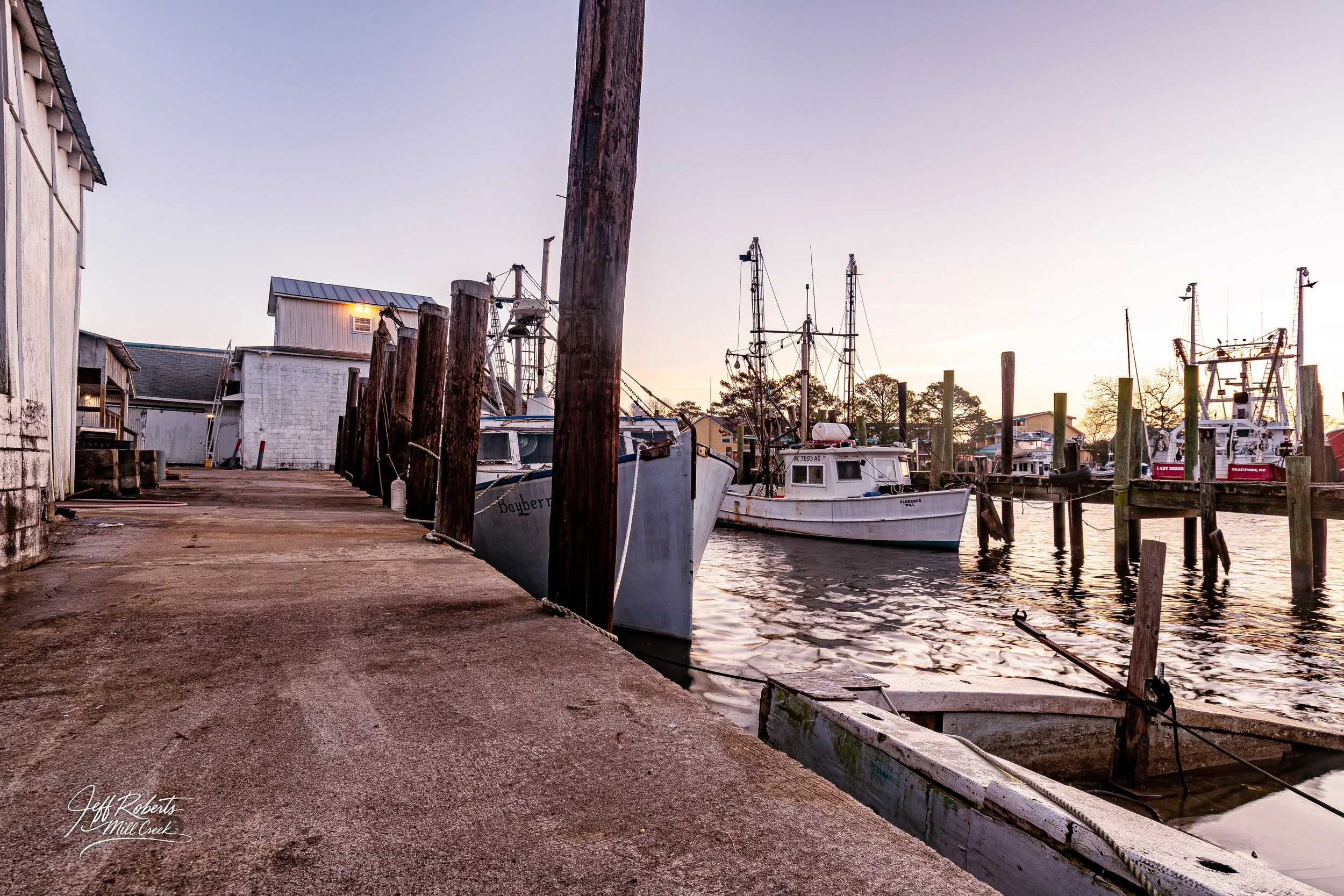 Dockside scene with boats moored at a marina at sunset, with wooden pilings and buildings in the background.