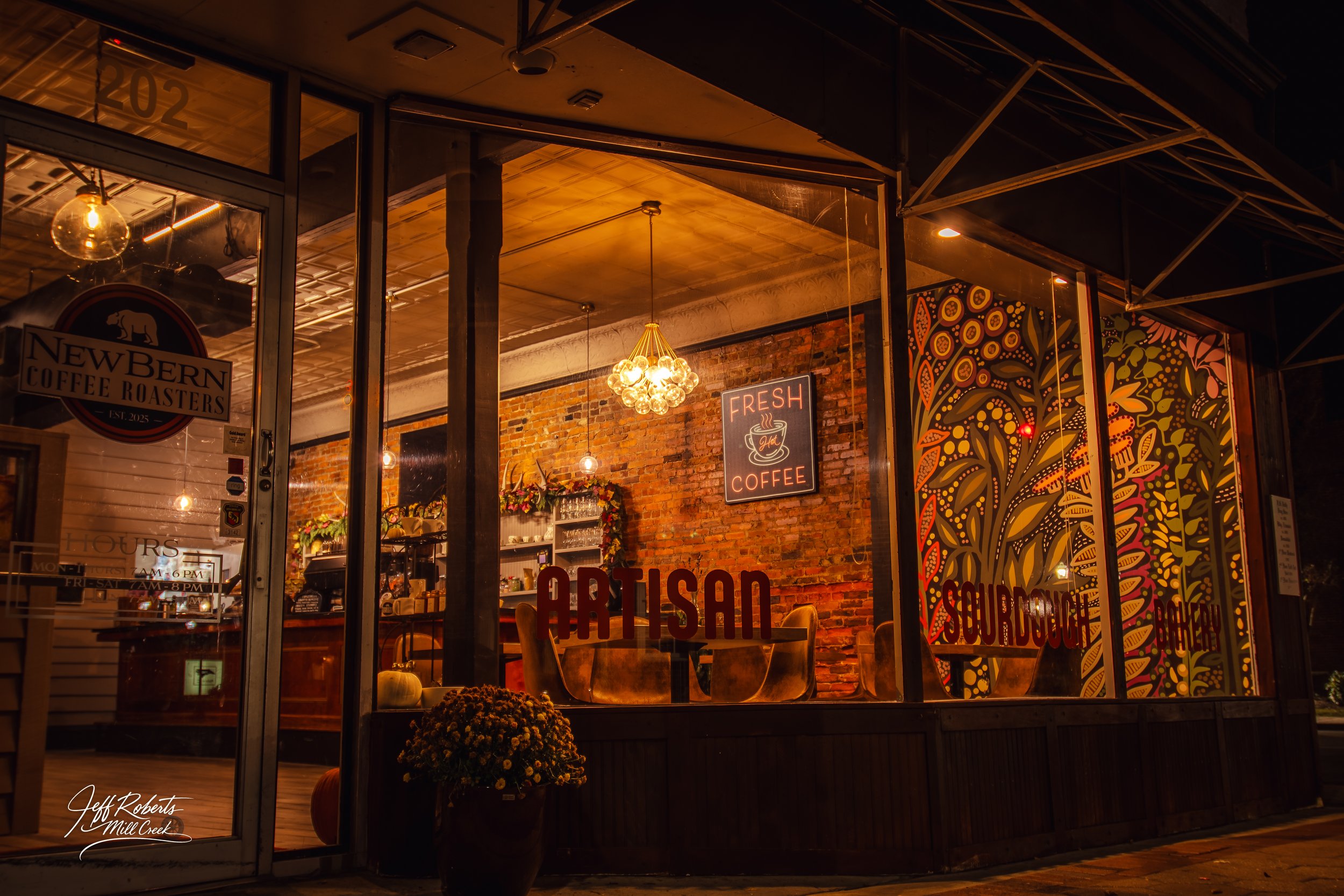 Exterior view of a cozy coffee shop at night with warm lighting, brick walls, colorful mural, and decorative signage displaying 'Fresh Coffee', 'Artisan', and 'Sourdough Bakery'