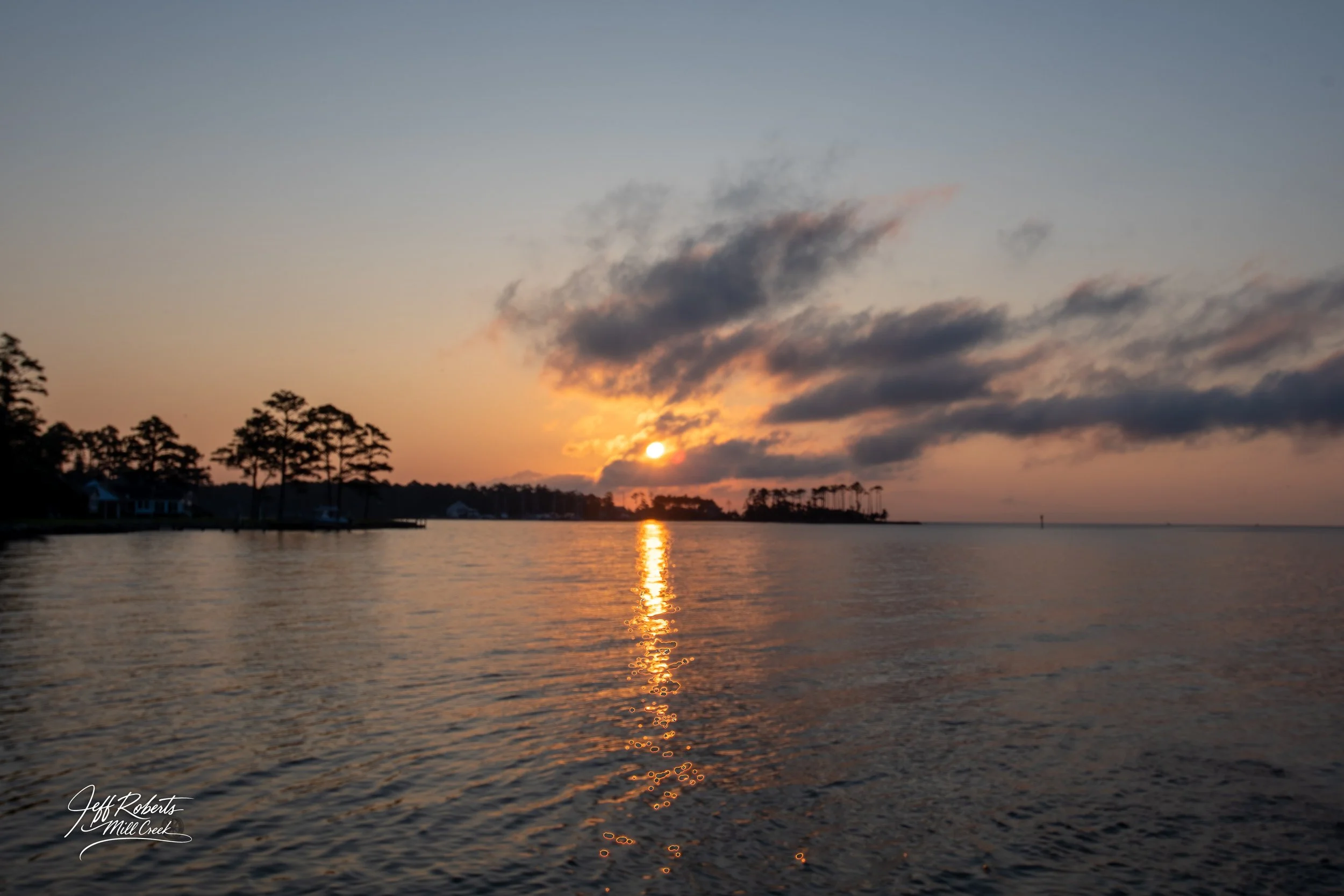 A sunset over a body of water with clouds in the sky and silhouettes of trees on the shoreline.