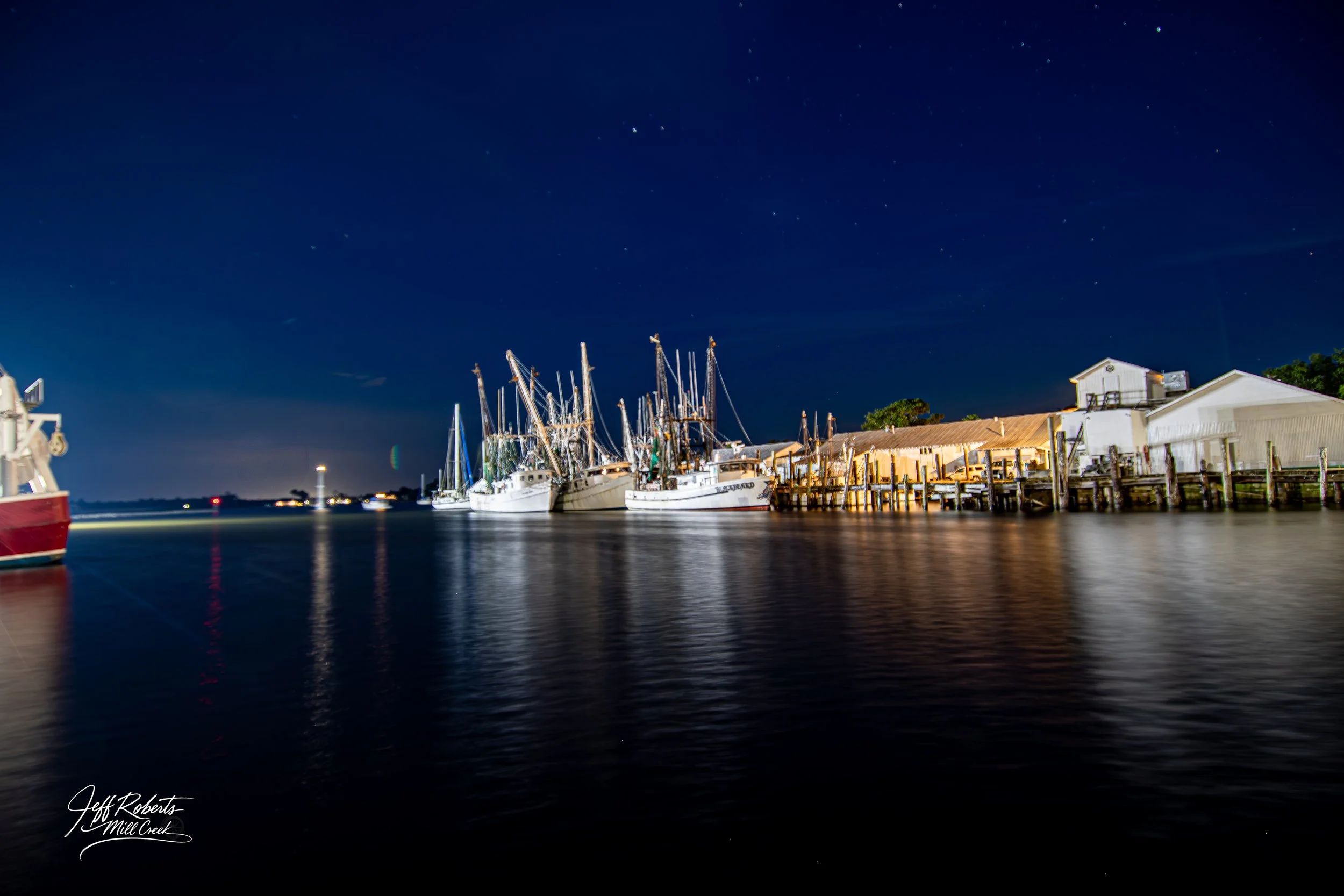 Nighttime view of a marina with several sailing boats docked, a wooden pier, and a large white building. The sky is dark with visible stars, and the water reflects the lights from the boats and buildings.