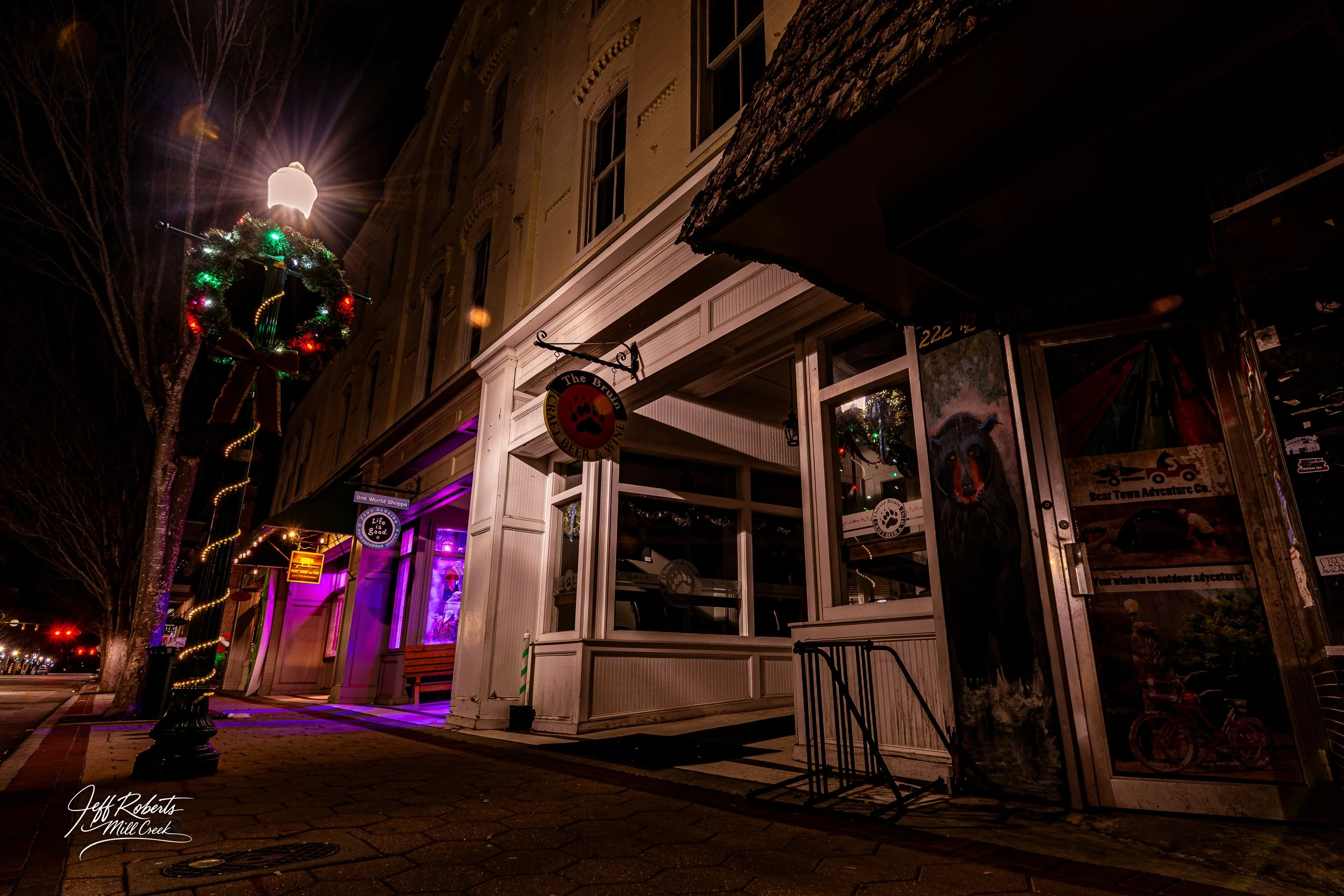 Night street view showing a storefront with a bear painting on the door, a sign for The Bramblebar, a lit Christmas wreath in the tree, and colorful lights inside the shop.