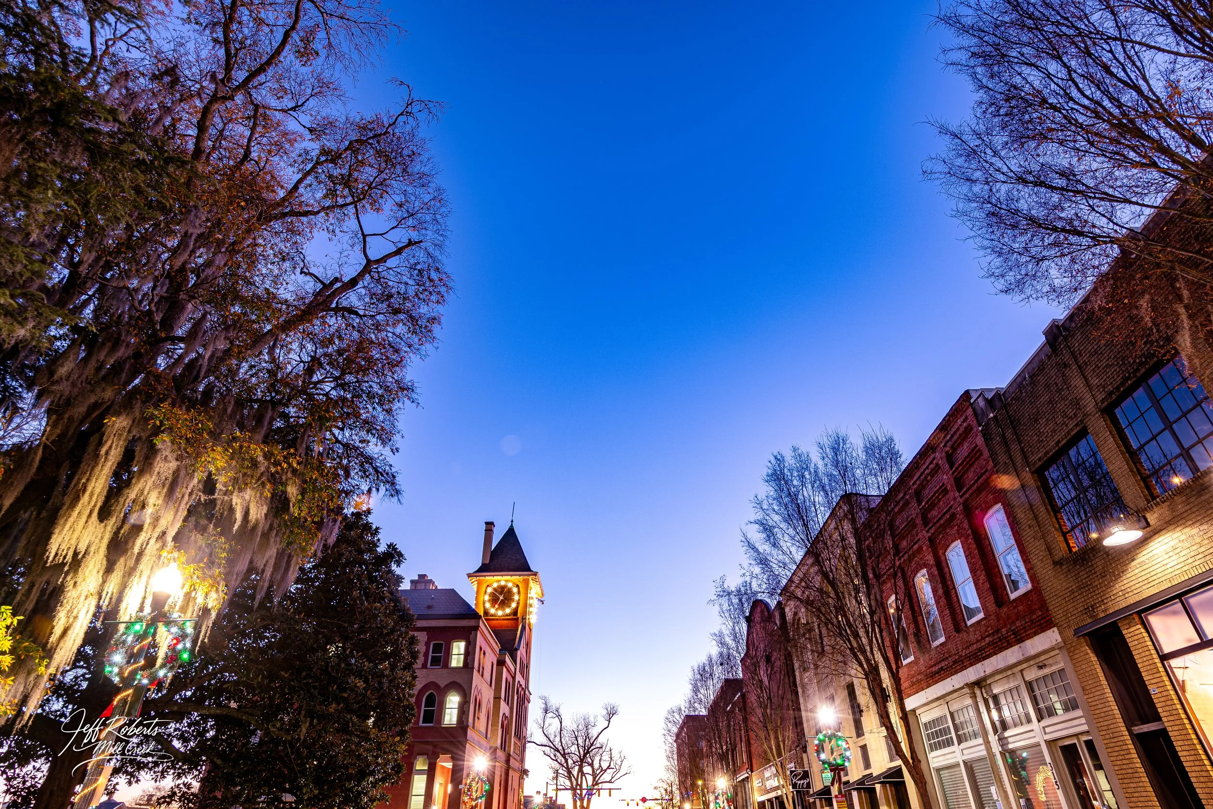 Dusk view of a small town with a clock tower, brick buildings, decorated Christmas wreaths, and trees with some Spanish moss hanging, under a clear evening sky.