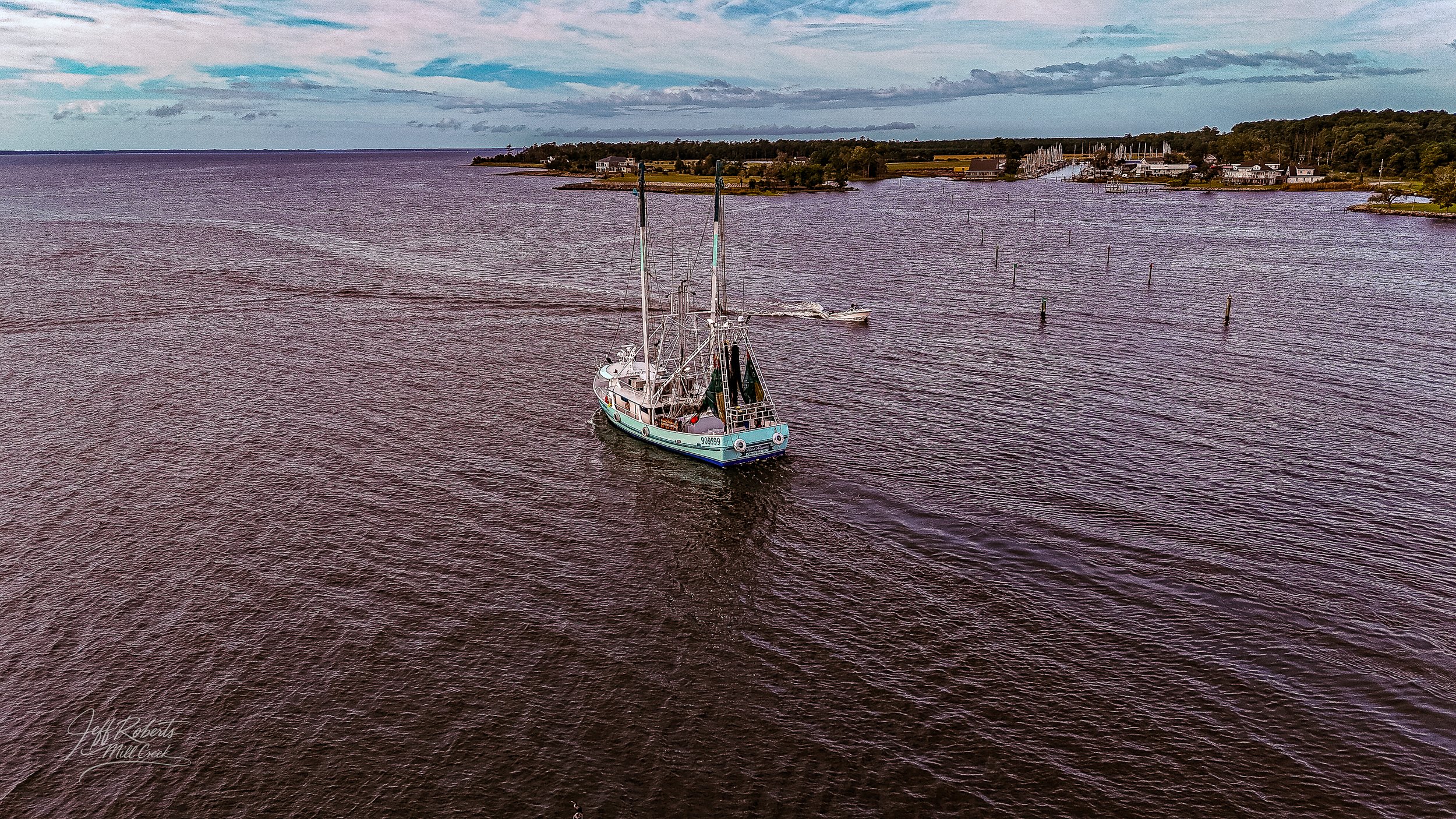 A sailboat with two tall masts sailing on a calm body of water near a shoreline with houses and trees, under a partly cloudy sky.