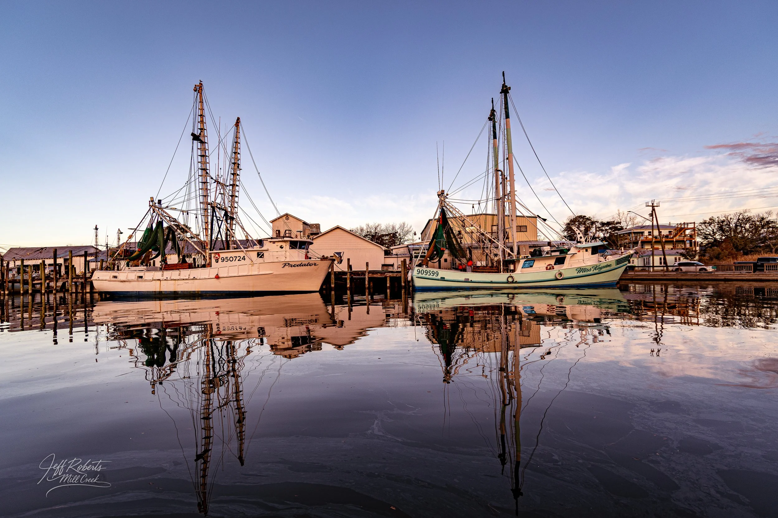 Two sailboats docked at a marina with their reflection visible in calm water, houses and trees in the background during sunset.