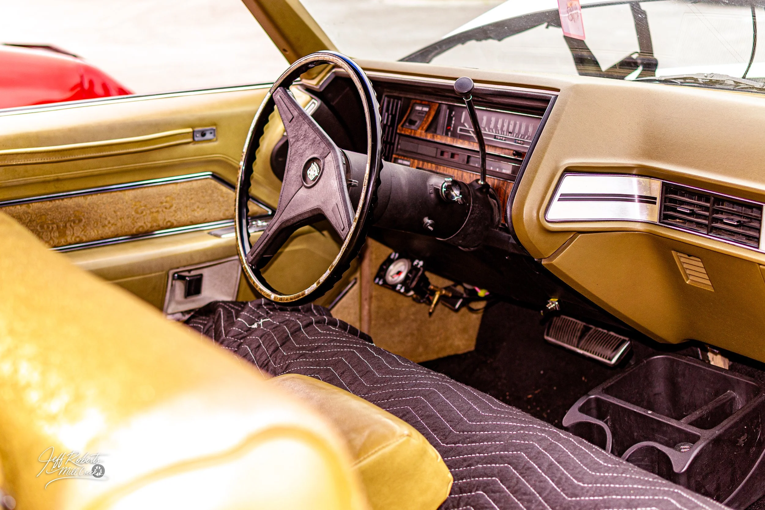 Interior of a vintage car showing a mustard yellow dashboard, steering wheel, and beige seats with a person sitting inside wearing dark pants with white stitching.