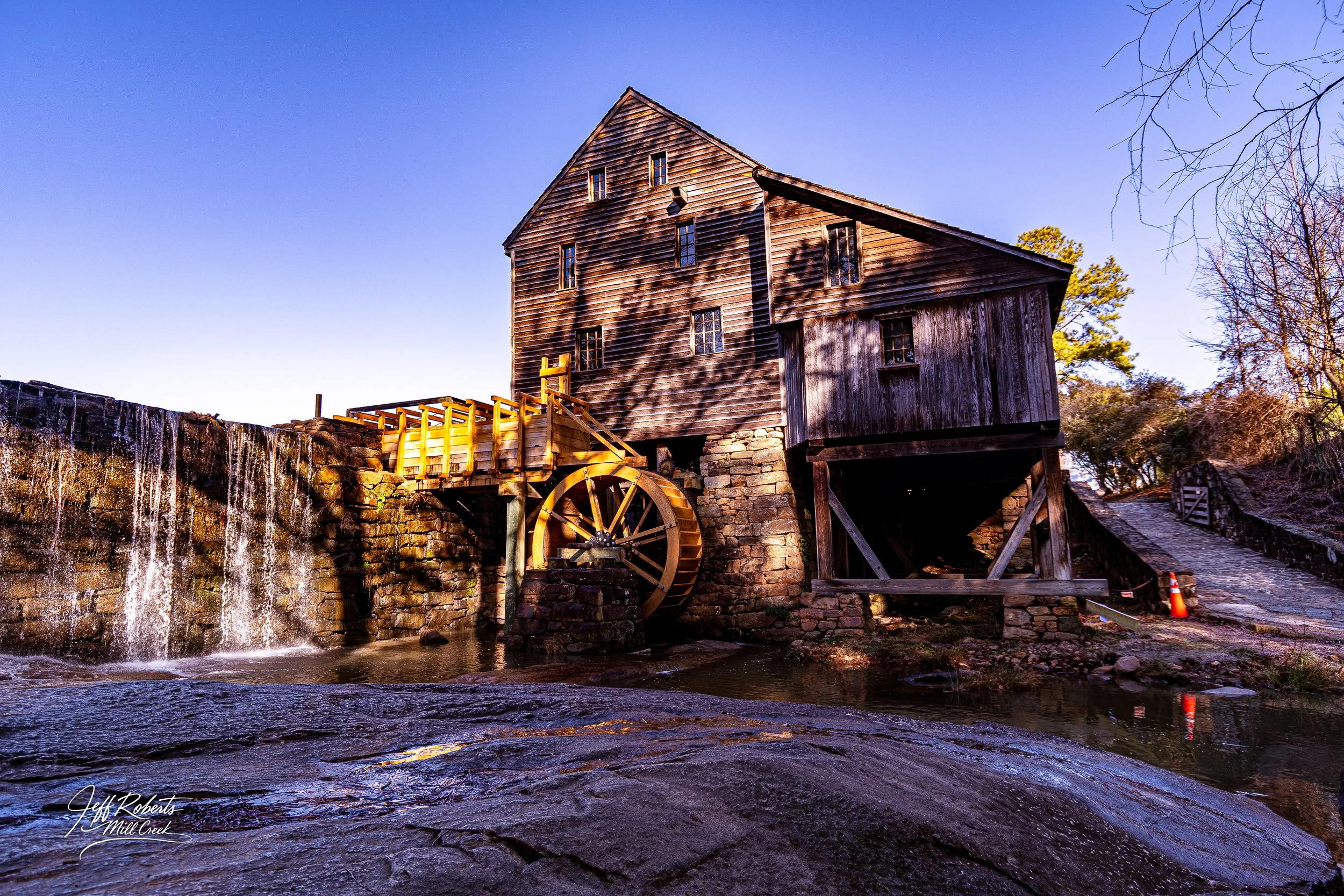 Old wooden water mill with a water wheel beside a flowing stream under a clear blue sky.