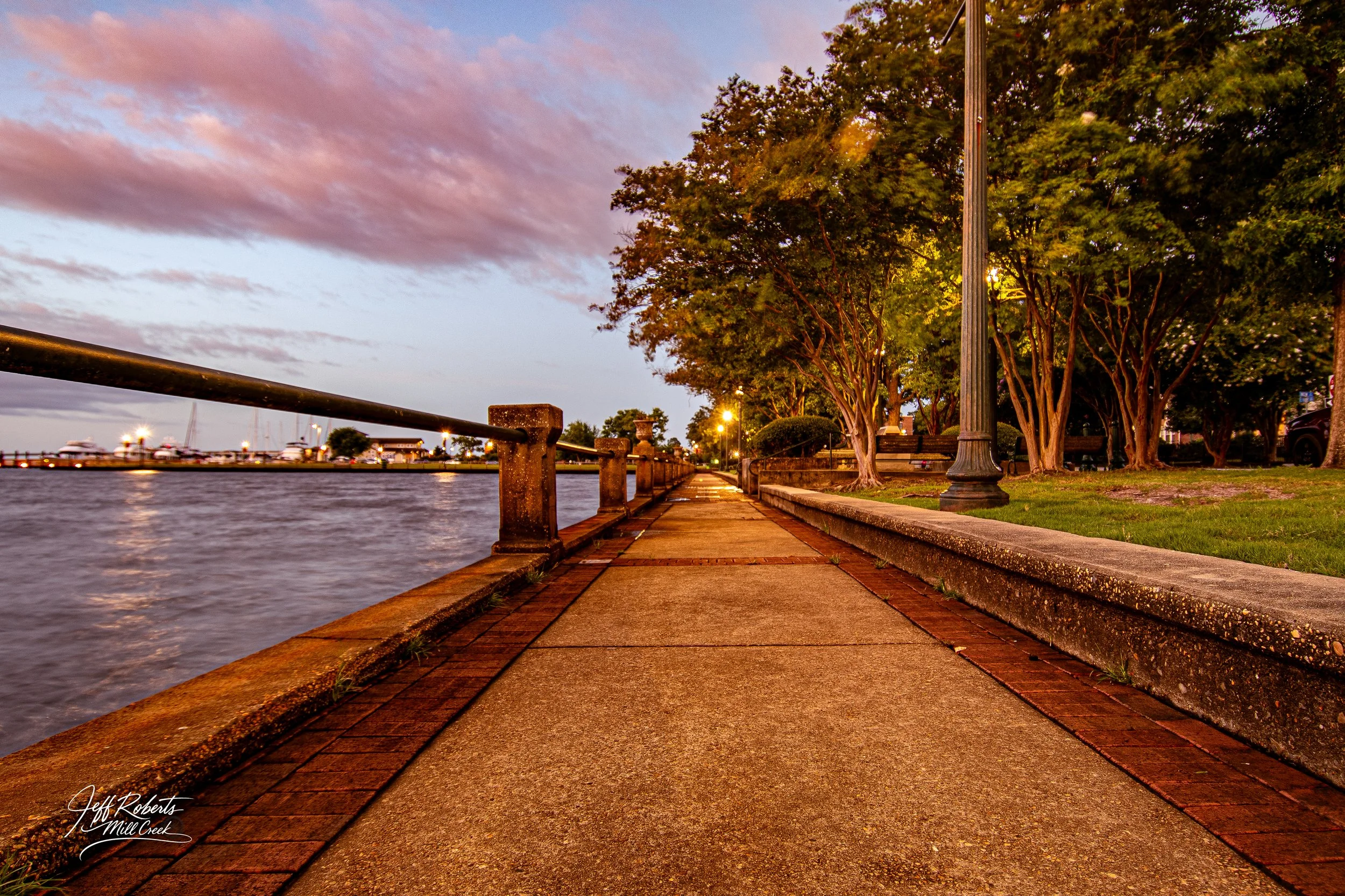A sunset view of a riverside walkway with trees, streetlights, and boats anchored on the water in the distance.