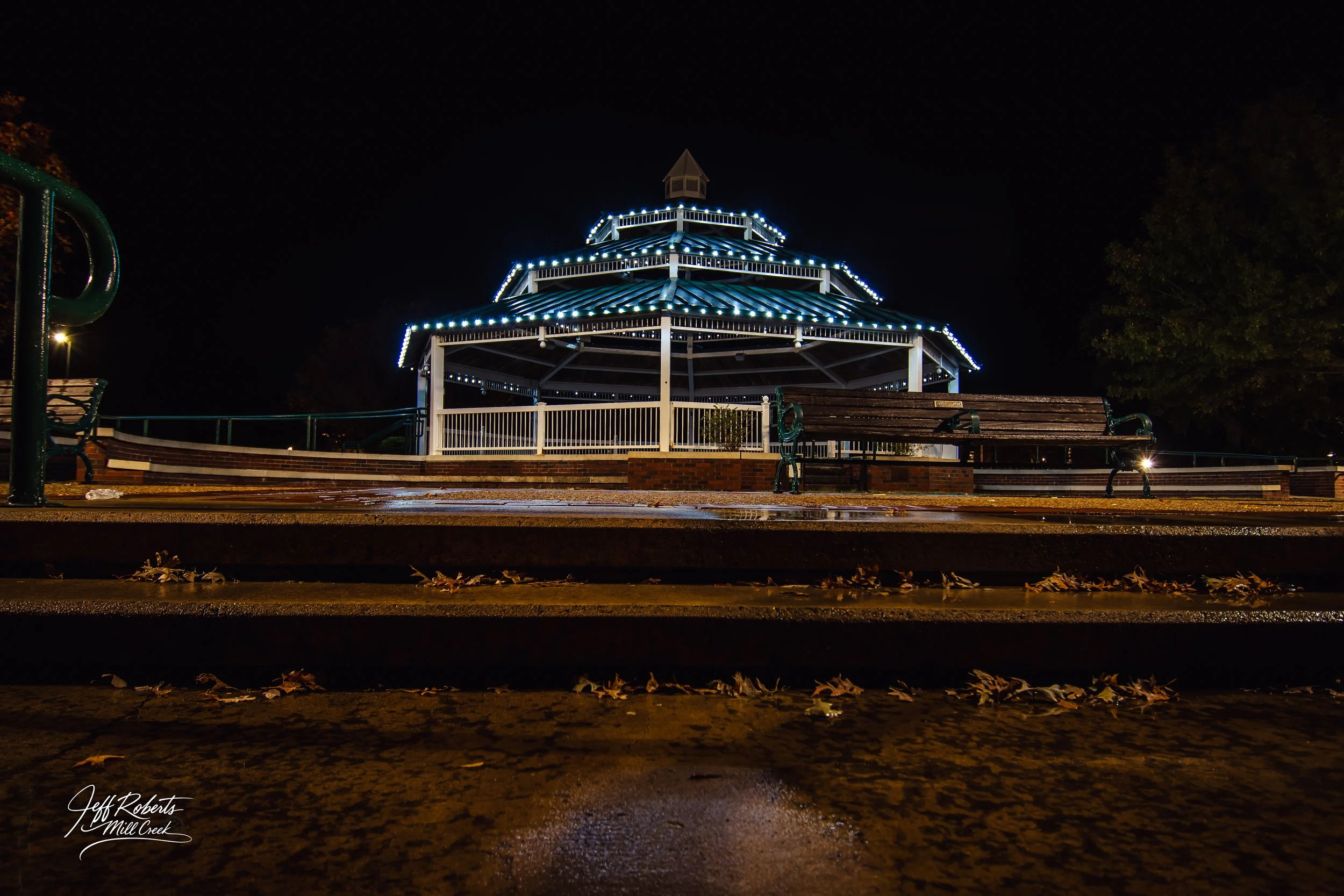 Night view of a lit-up pavilion with benches, surrounded by trees, and steps with fallen leaves in the foreground.