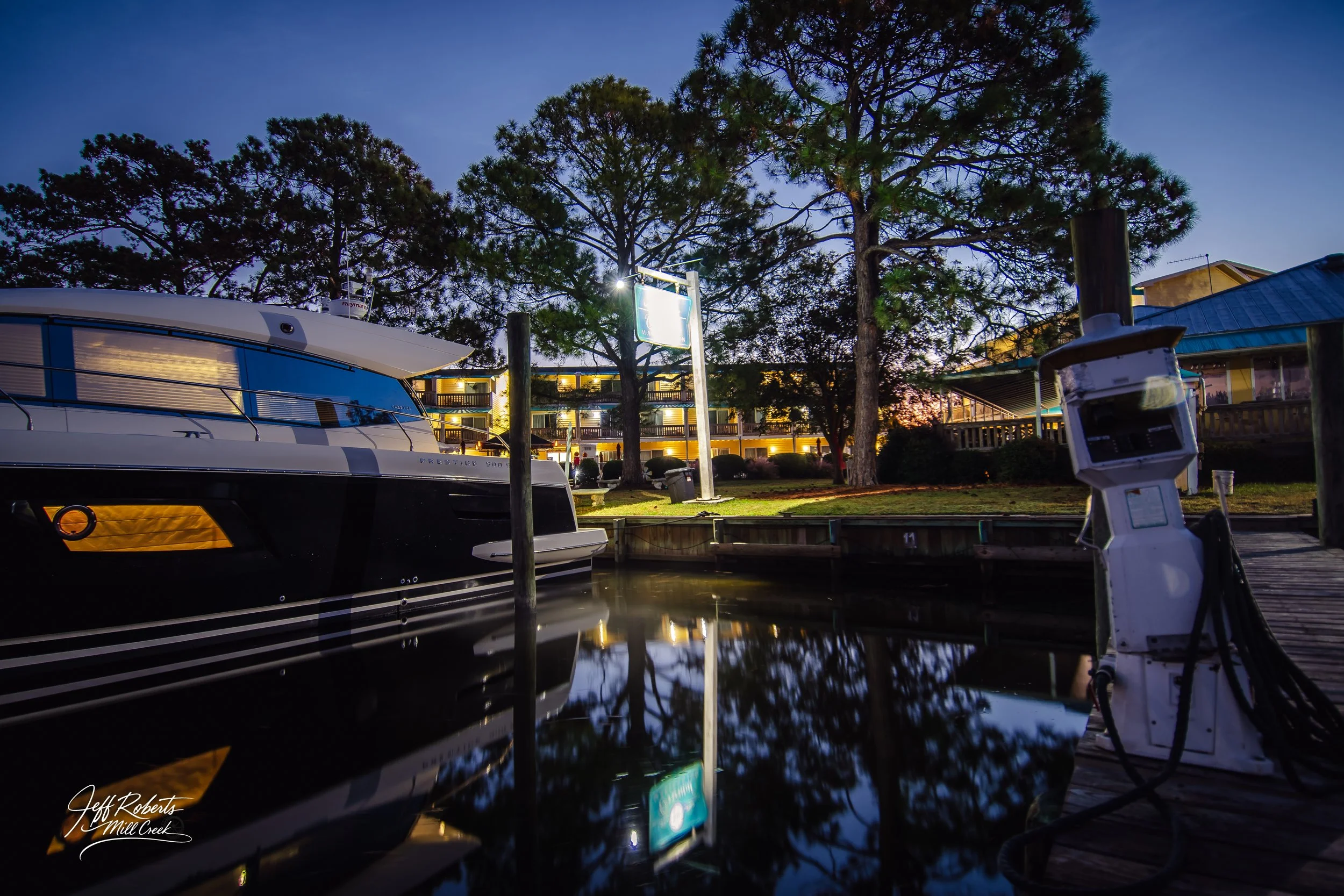 A marina at dusk with a luxury yacht docked, trees in the background, separate buildings on the right, and an electric boat charger in the foreground, reflecting on calm water.