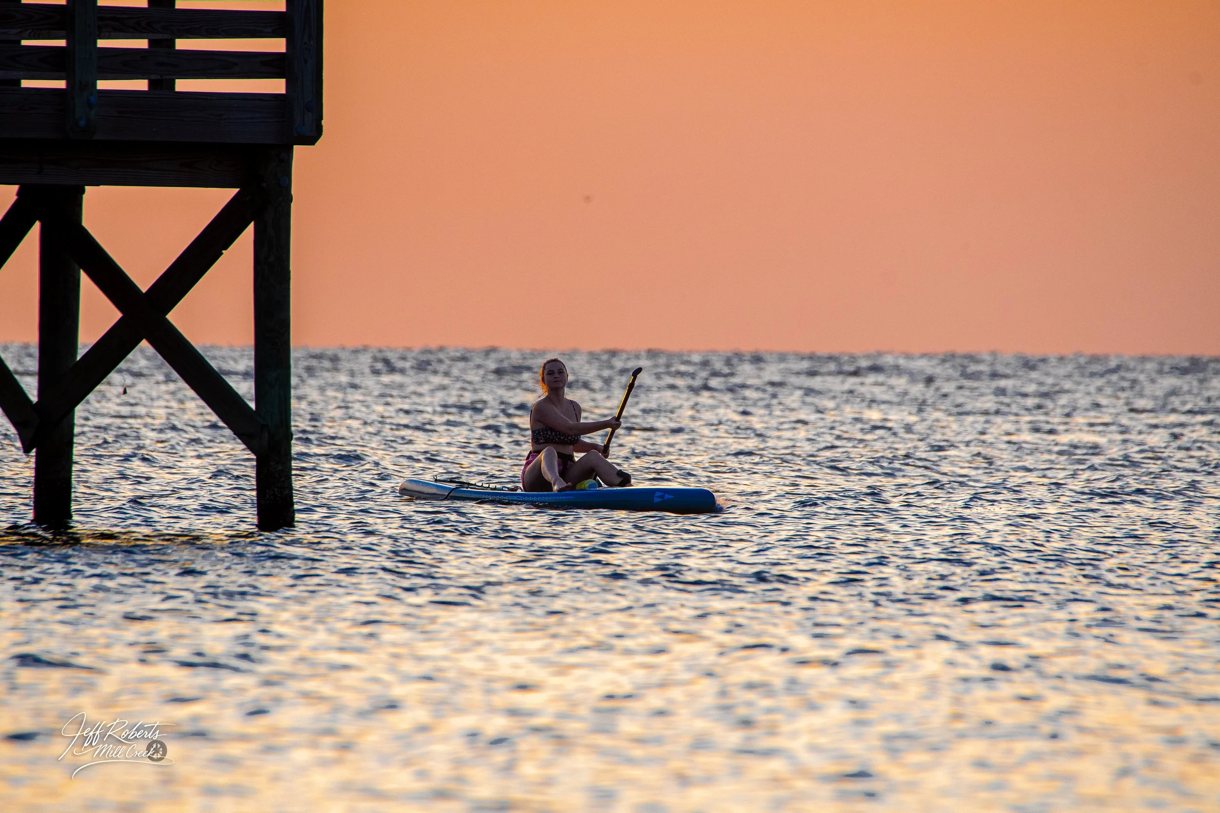 Person paddleboarding near a wooden pier during sunset with a pink sky.