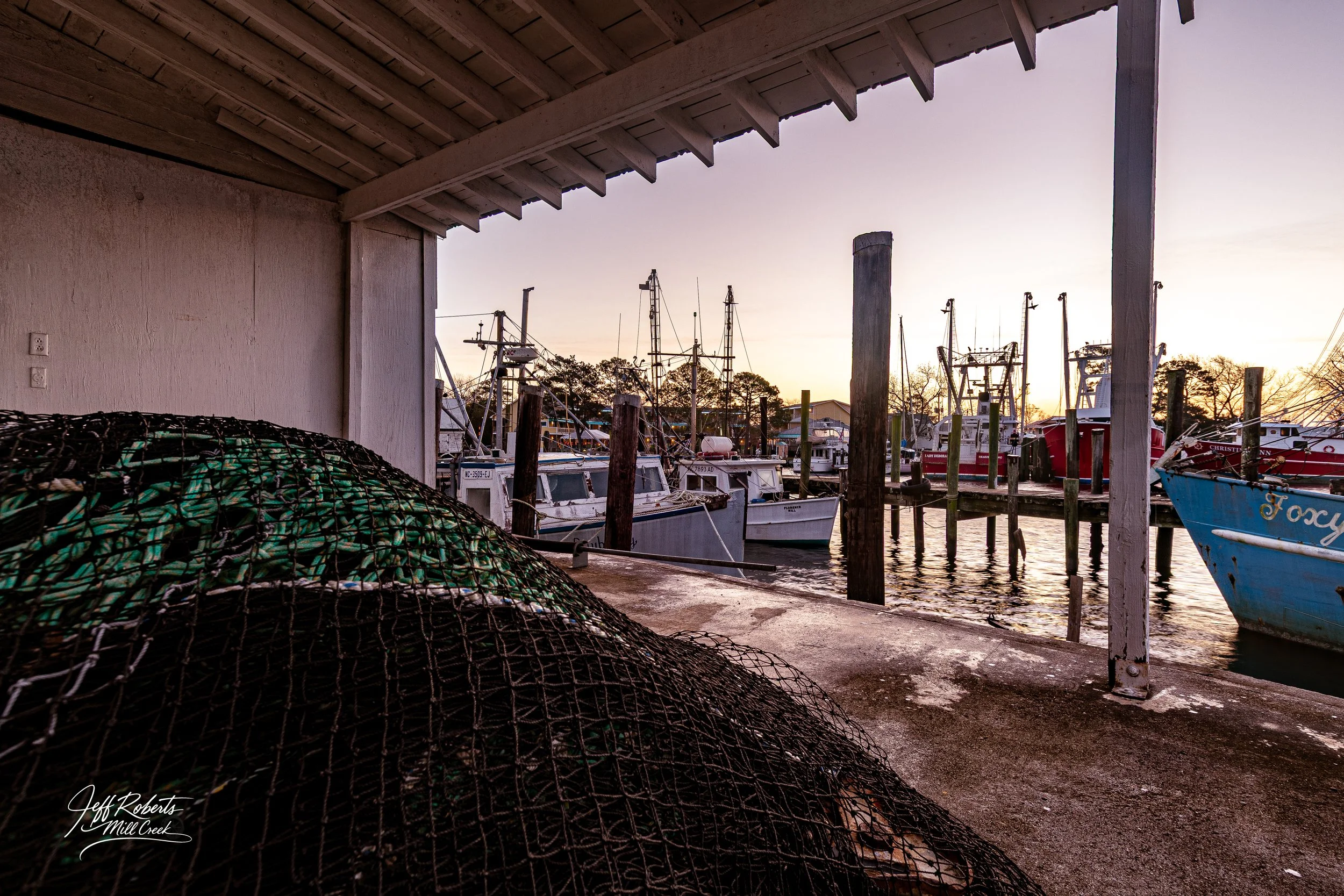 View of boats docked at a marina during sunset, with fishing nets in the foreground and wooden dock posts.