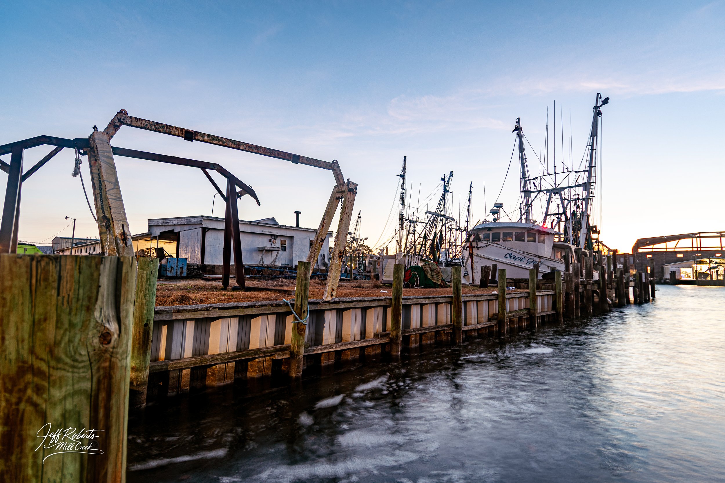 Docked boats along a canal with a small white building, some rusted equipment, and a bridge in the background at sunset.