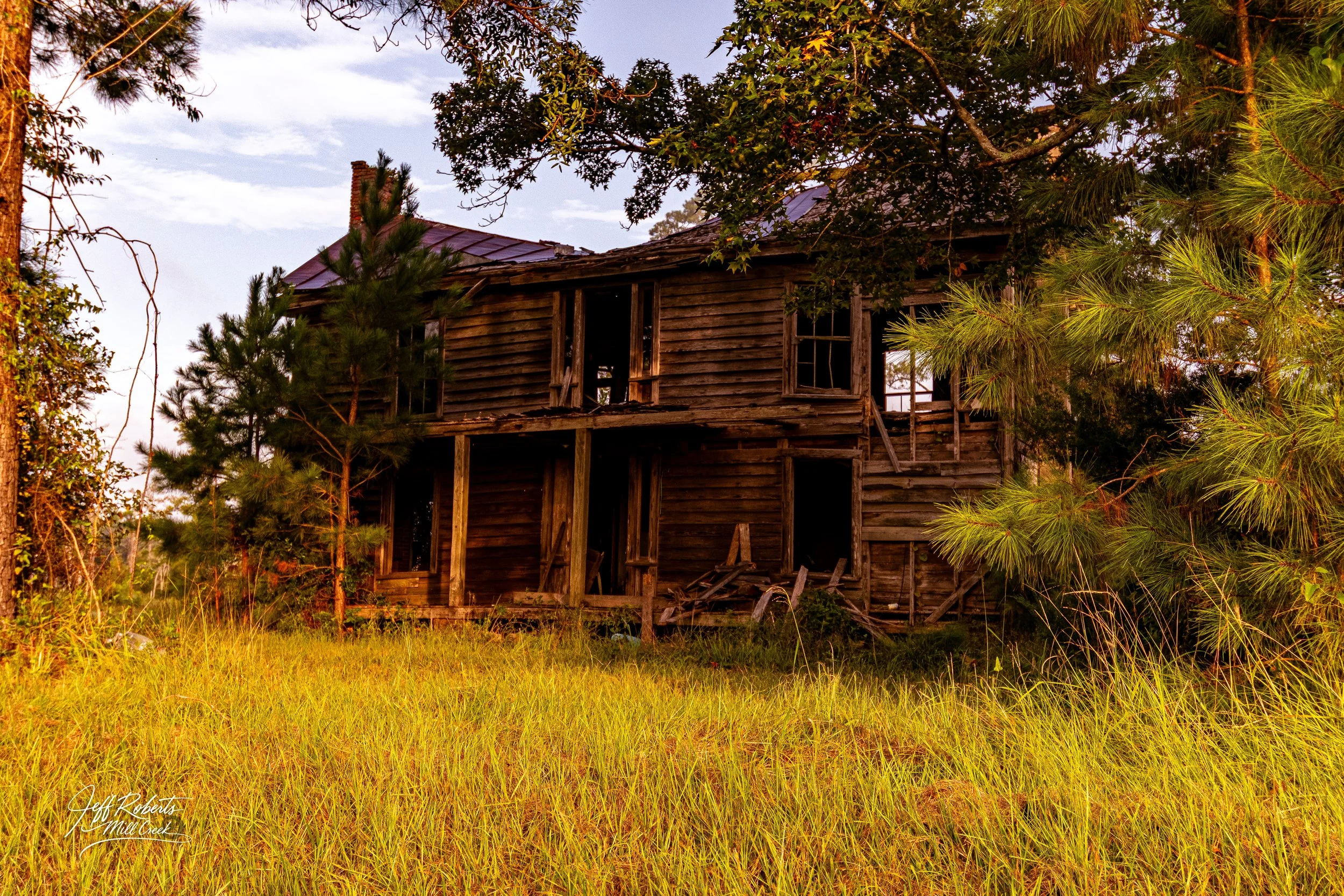 An abandoned, dilapidated two-story wooden house surrounded by overgrown grass and trees, with broken windows and missing roof sections, under a partly cloudy sky.