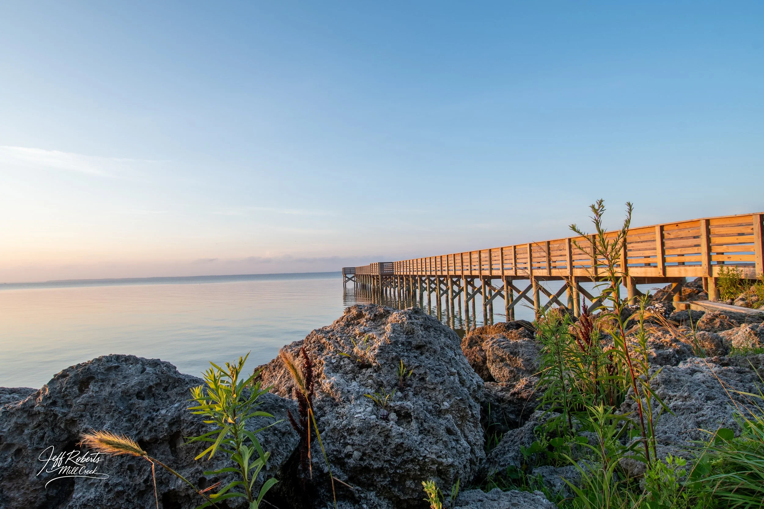Wooden pier extending over rocks into calm water at sunset or sunrise with clear sky.