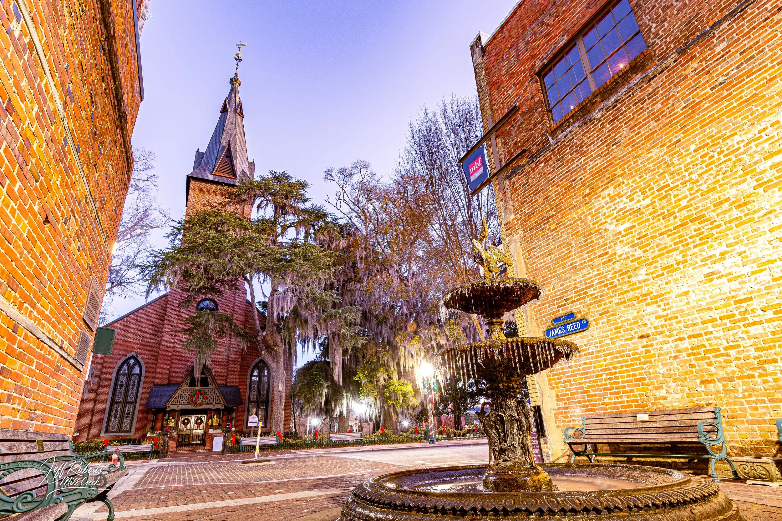 A vintage fountain with a gold sculpture on top is surrounded by brick buildings and a church with a steeple, trees, and Christmas decorations in a town square at dusk.