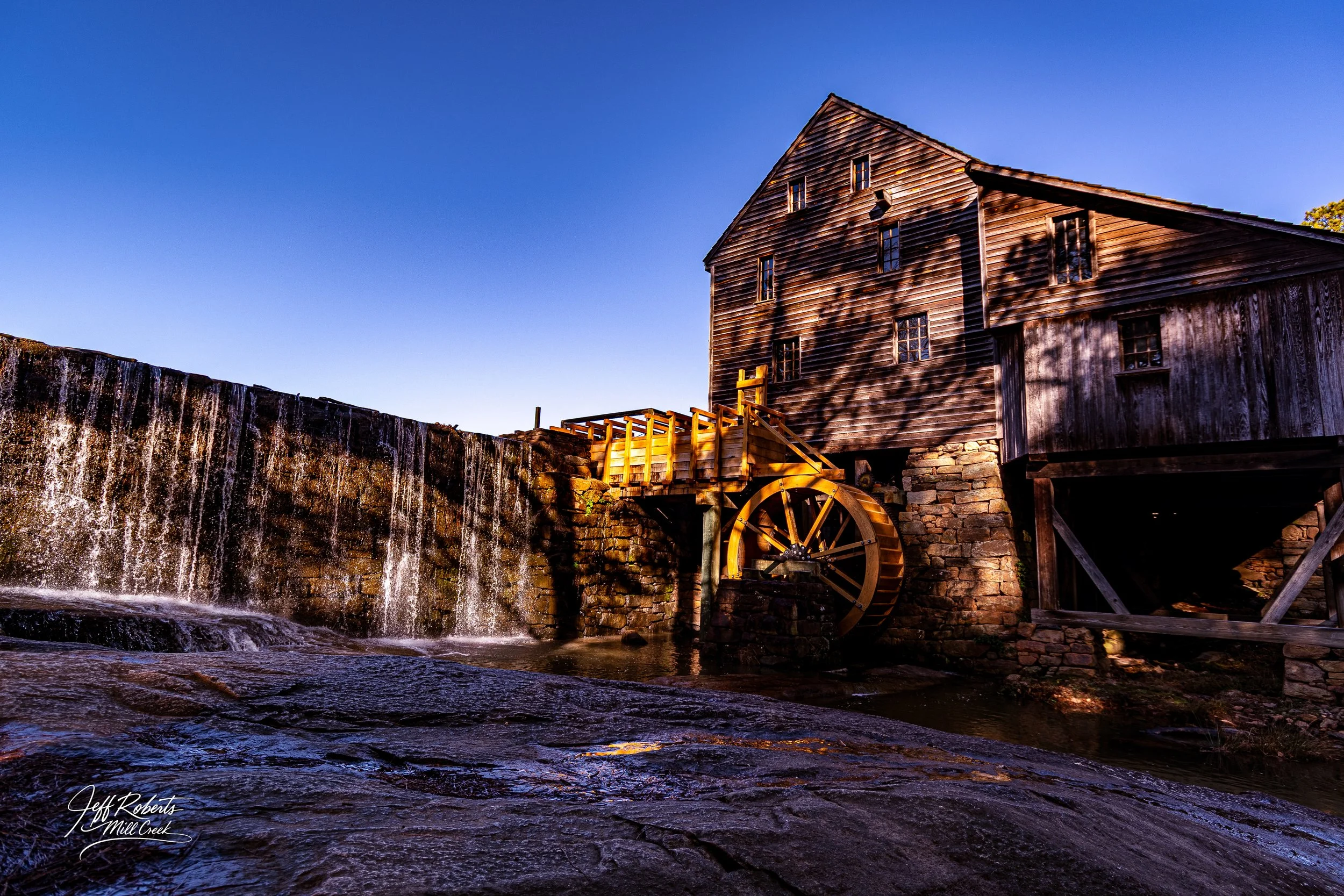 Old wooden water mill with a water wheel beside a waterfall under a clear blue sky.