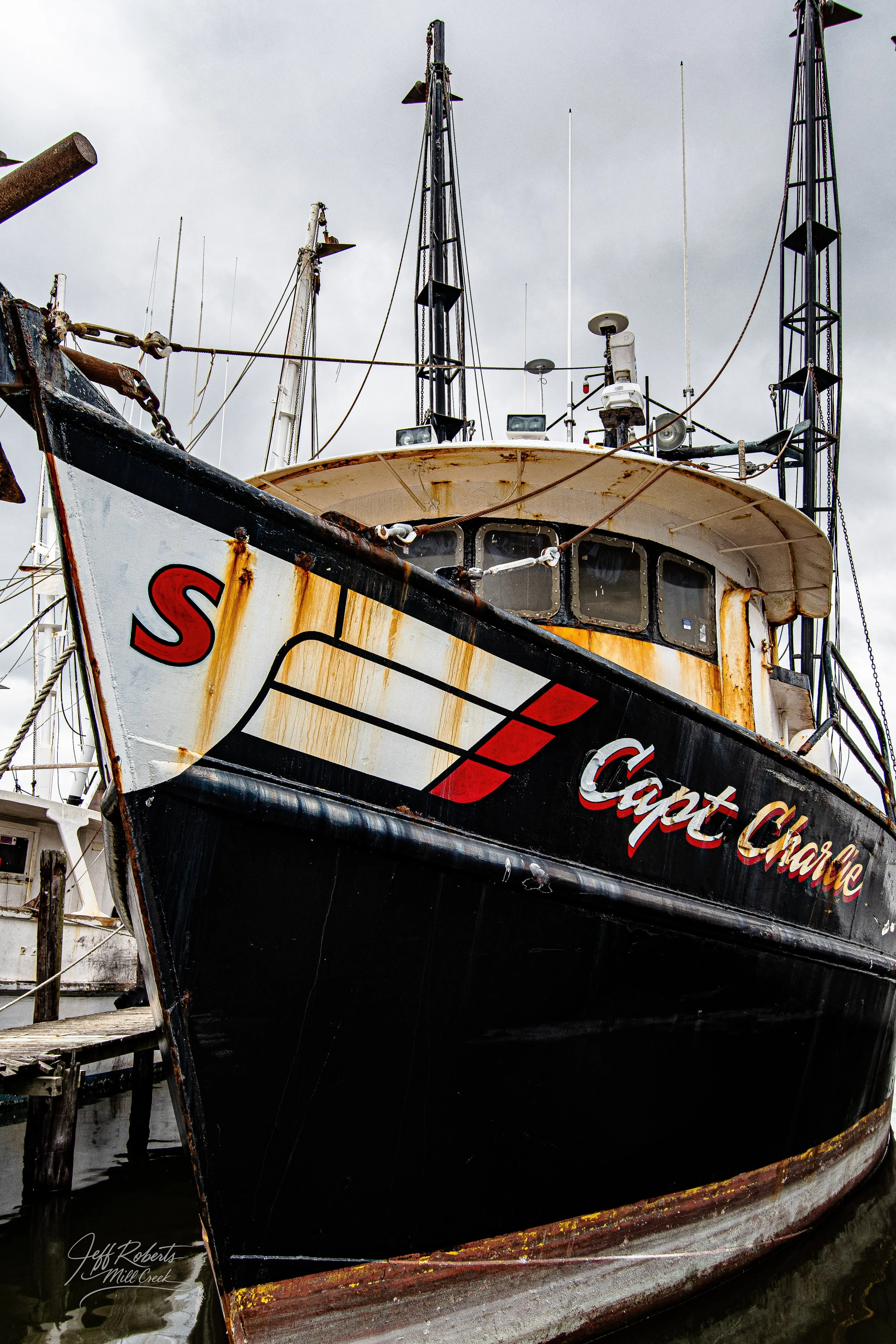 Close-up of a rusty, black and white fishing boat named 'Capt Charlie' docked at a pier with an overcast sky.