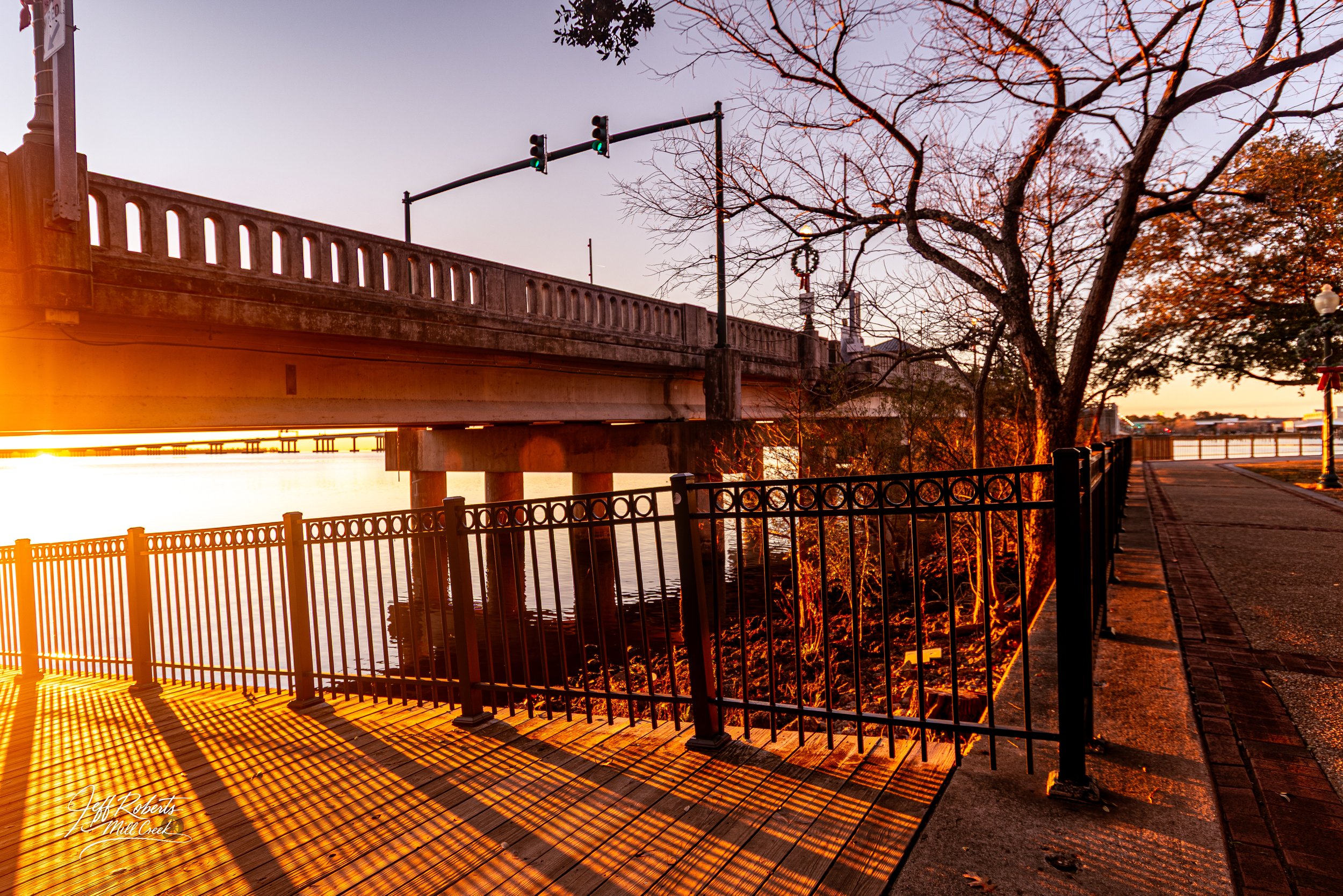 Sunrise over a bridge along a river, with trees and a paved walkway with a black metal fence and shadows cast on the ground.