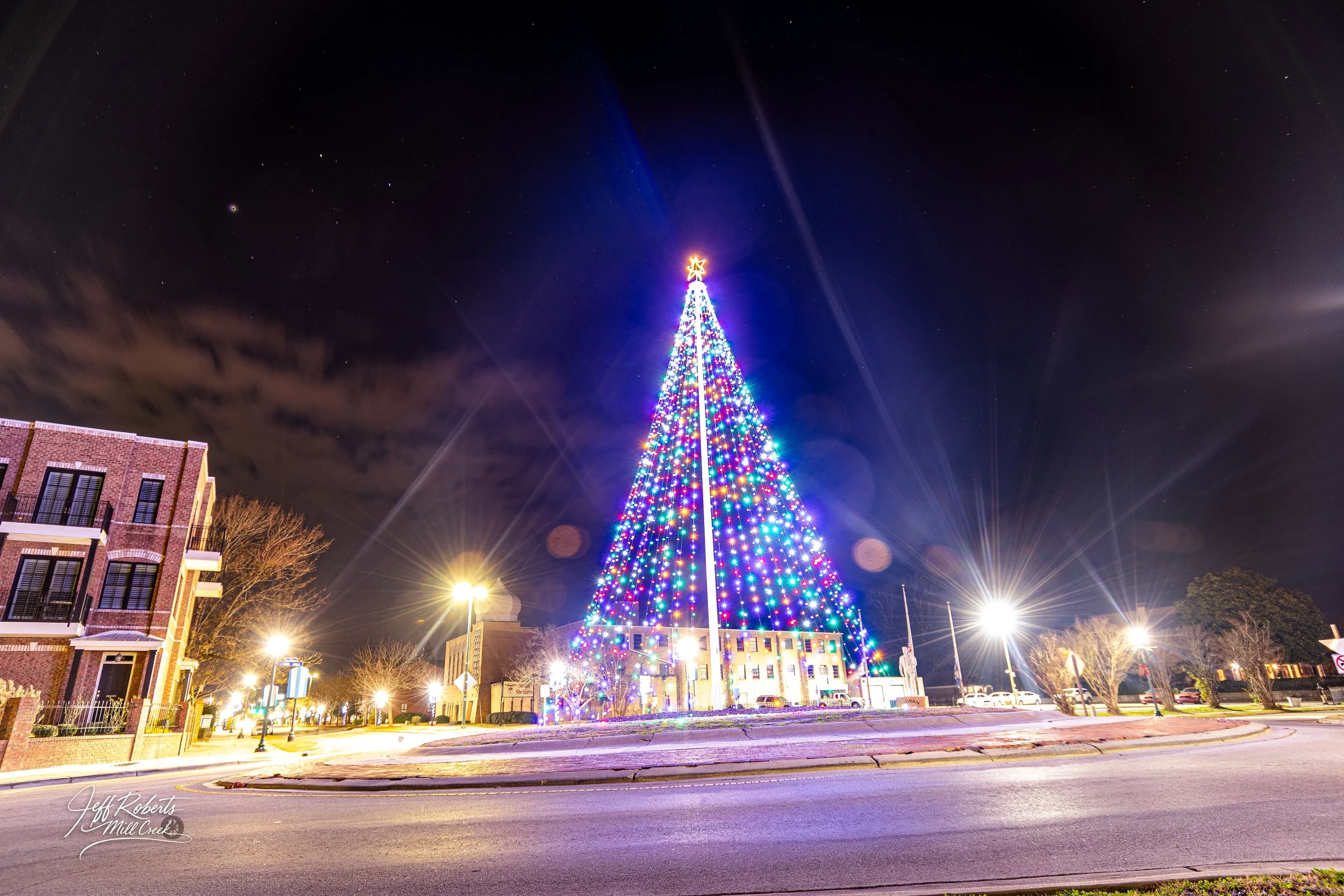Nighttime view of a large Christmas tree decorated with multicolored lights and topped with a star, surrounded by a small town square with streetlights, buildings, and trees.