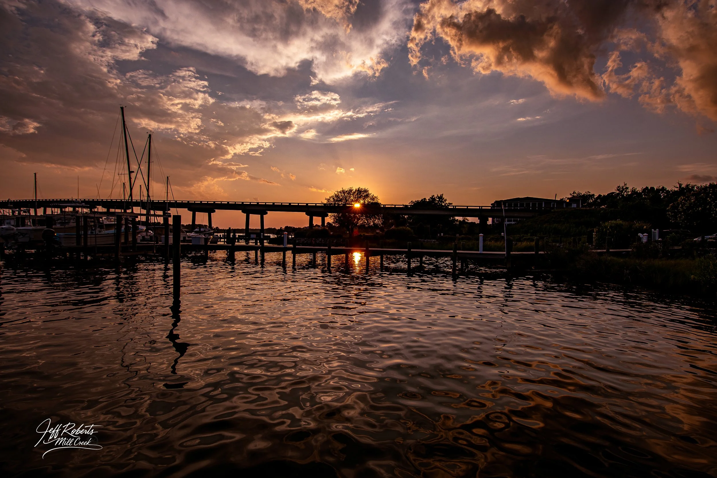 Sunset over a harbor with boats and docked piers, a bridge in the background, partly cloudy sky, and water reflecting the sky and sunlight.