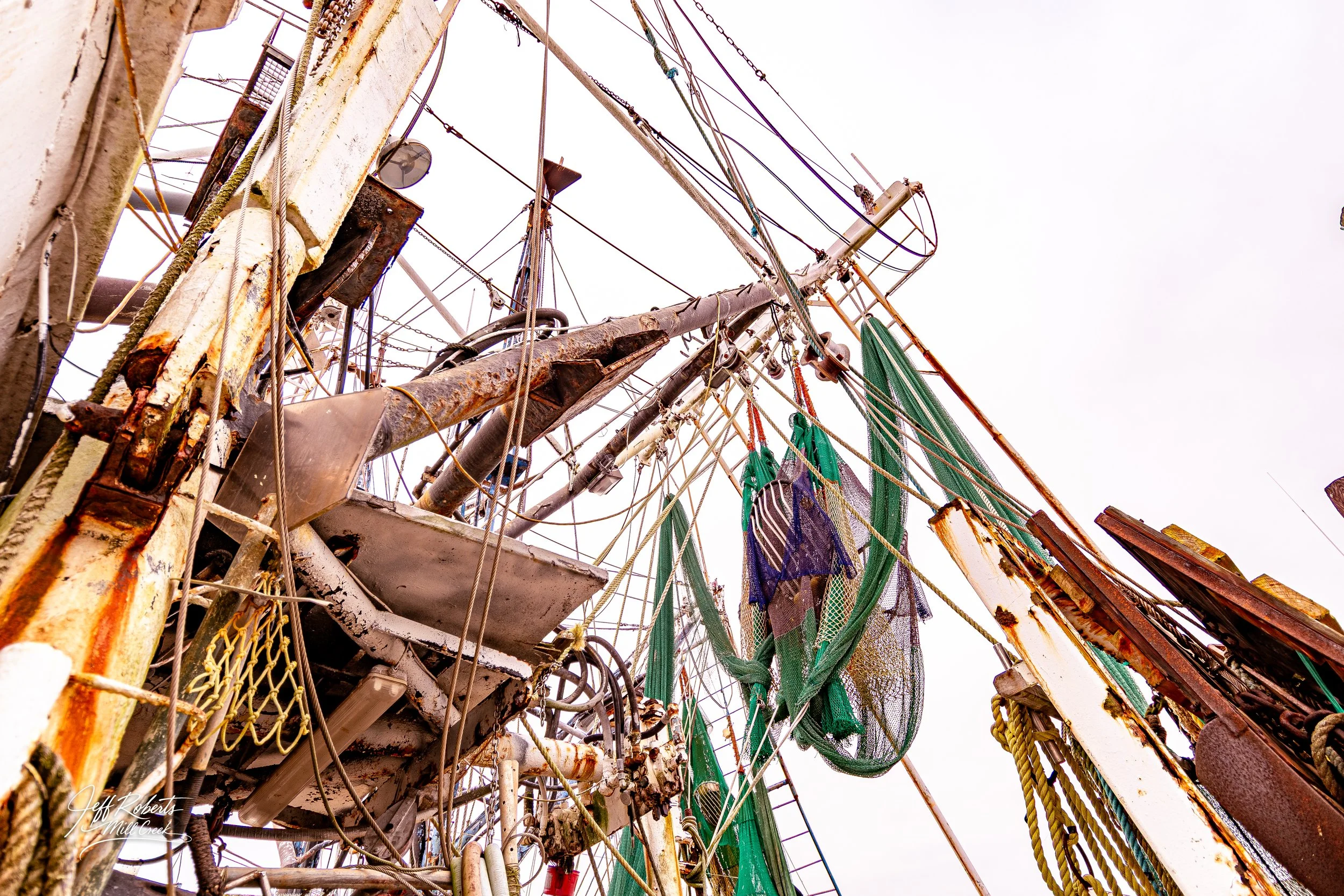 A view of a rusty, old pirate ship's mast with rigging, nets, and ropes against a cloudy sky.
