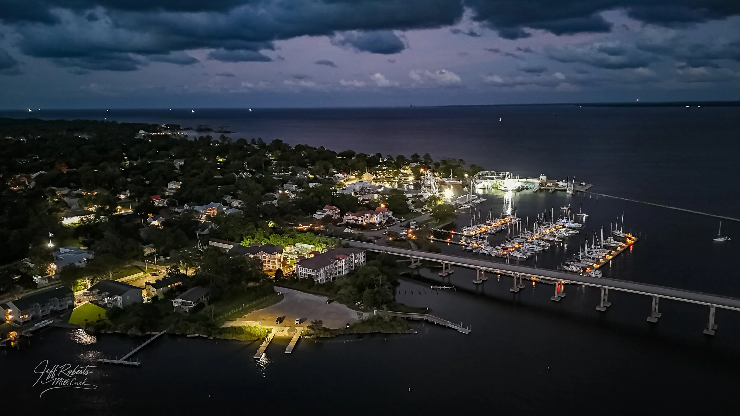 Nighttime aerial view of a marina with sailboats docked, illuminated buildings, a bridge, and a residential area near a large body of water under dark, cloudy skies.
