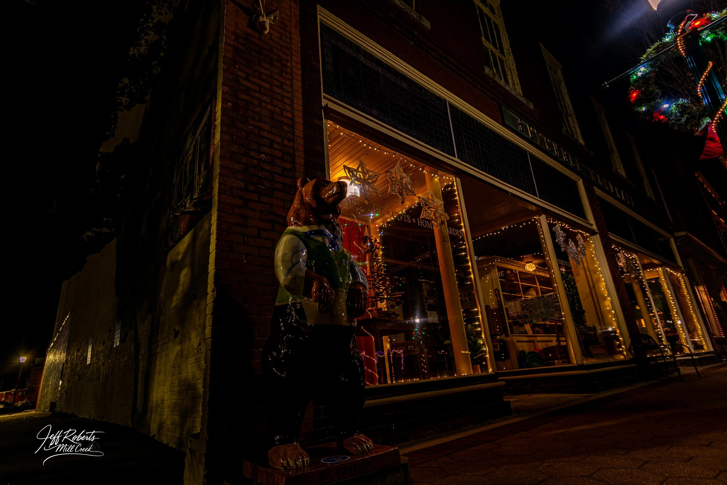 Nighttime view of a storefront decorated with festive lights and ornaments, with a bear statue wearing a jacket standing outside.