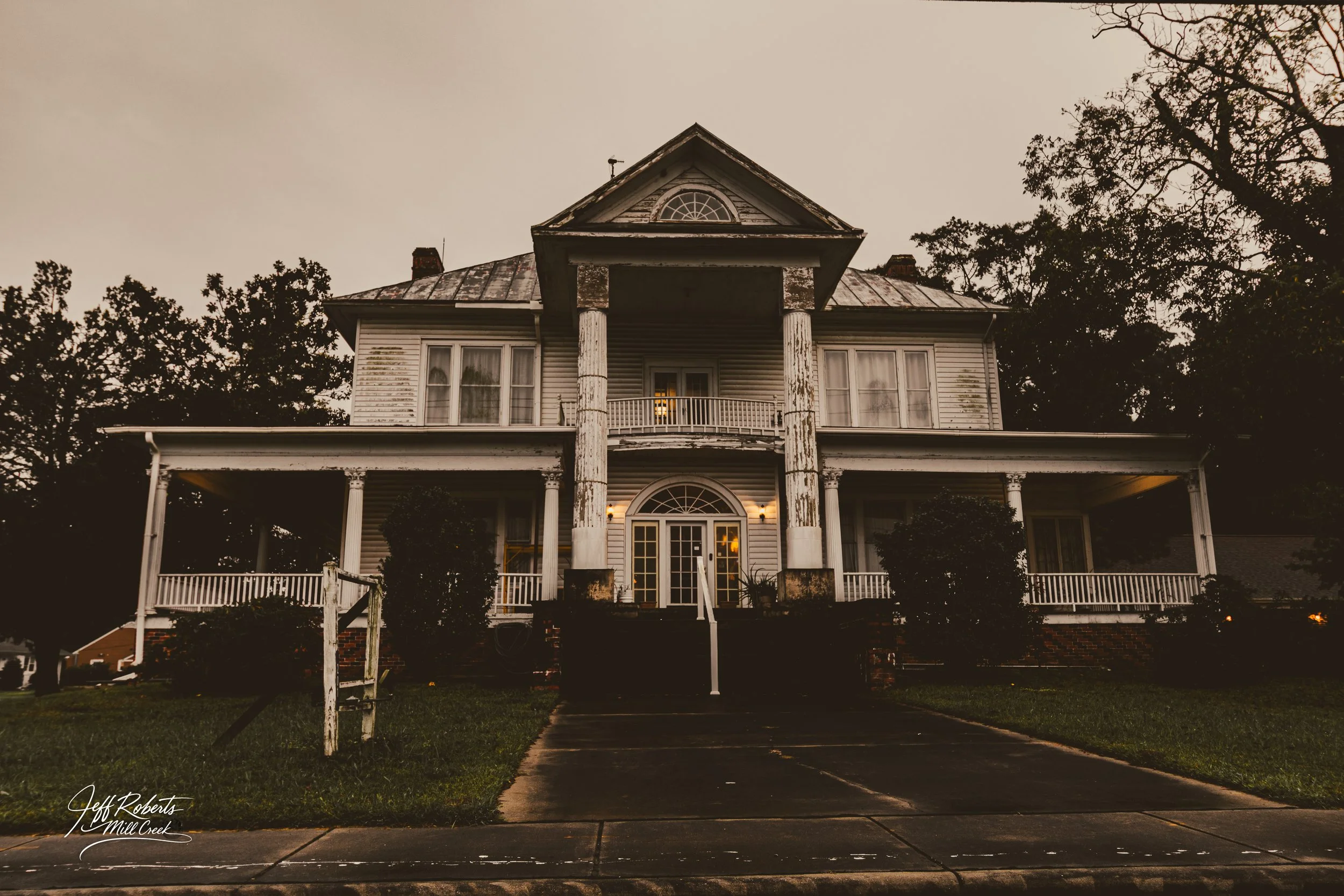 An old, large, two-story house with a porch and columns, showing signs of wear and tear, with trees surrounding it, during dusk.