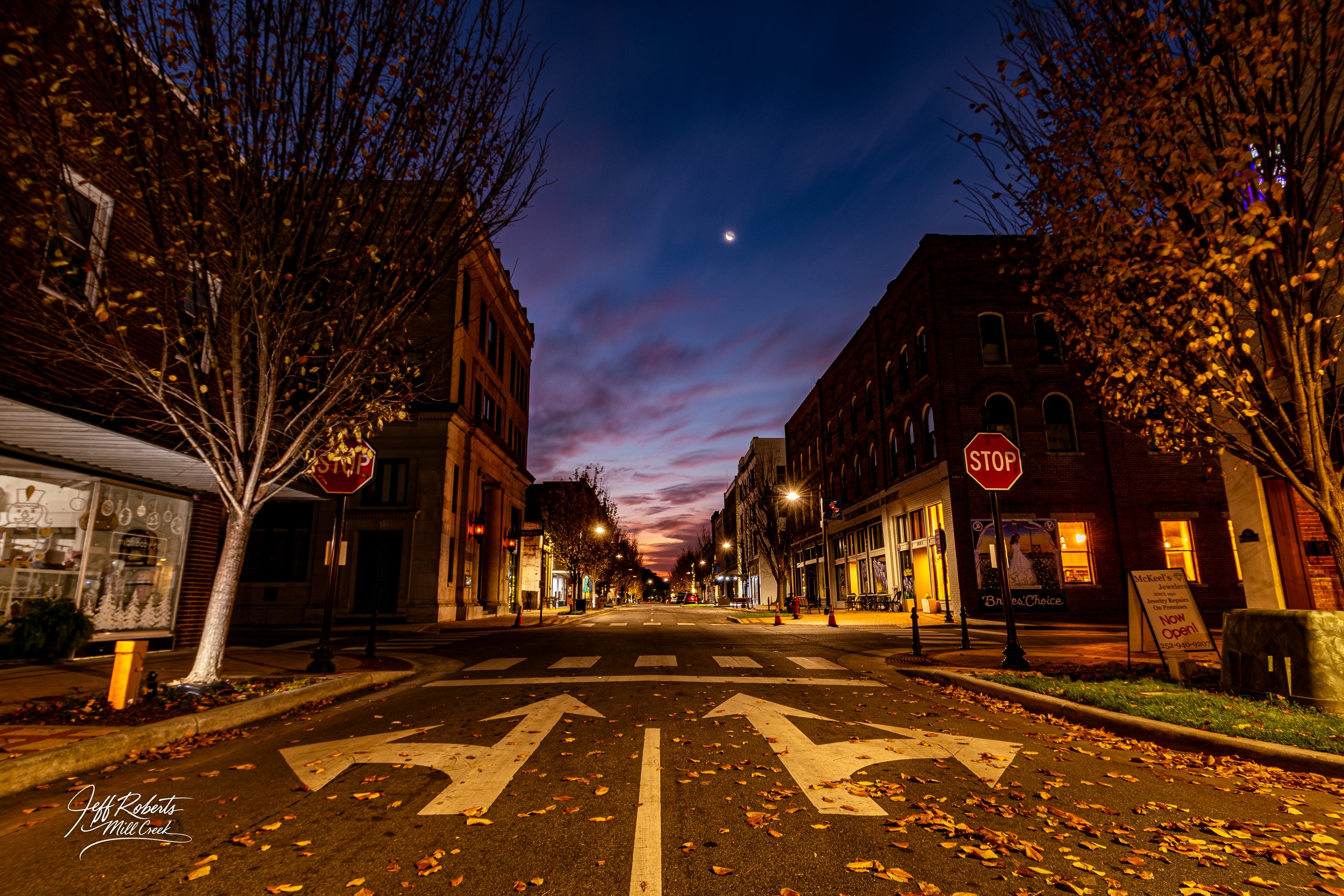 A quiet city street at dusk with falling autumn leaves, illuminated storefronts, streetlights, stop signs, and a crescent moon in the clear sky.