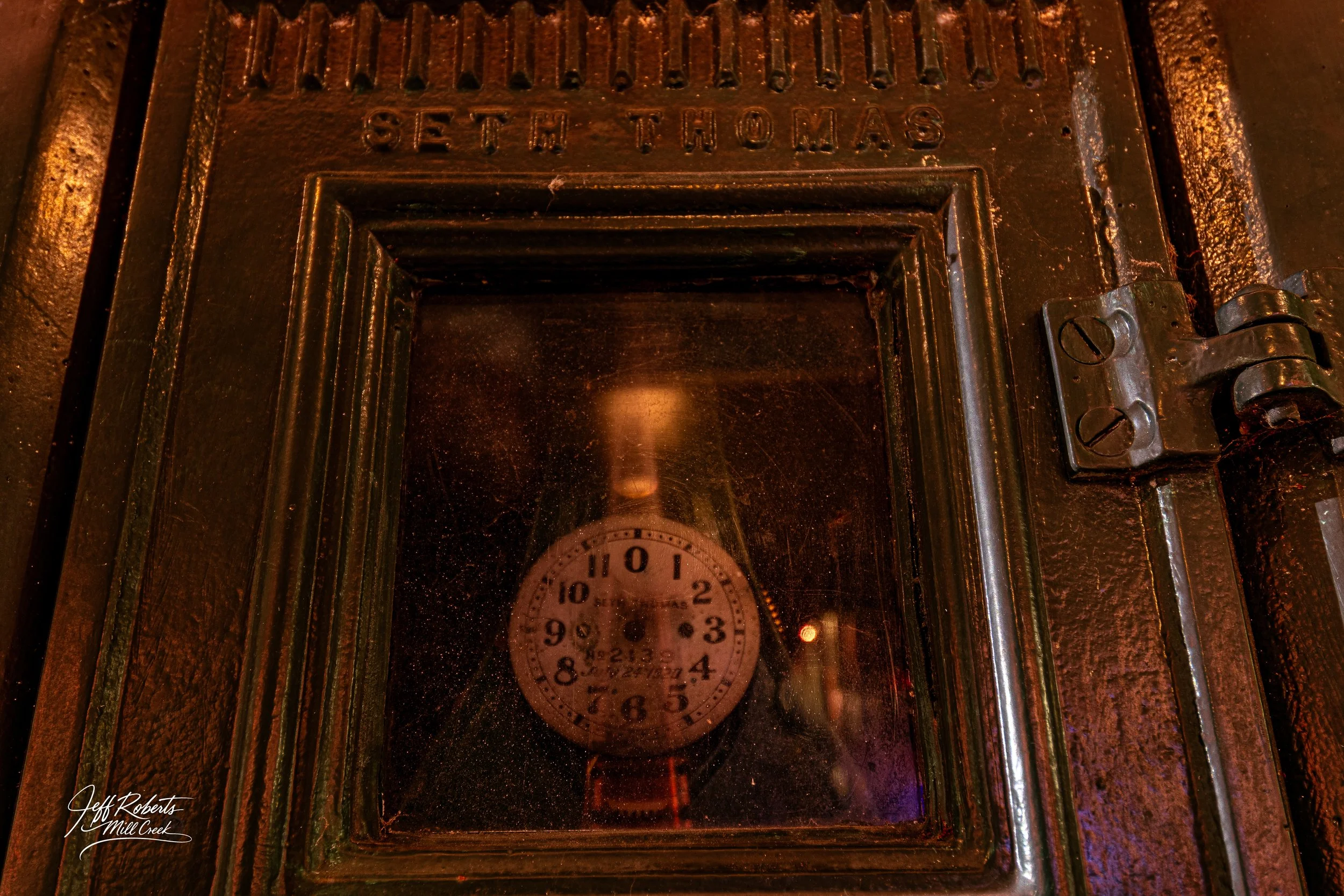 An antique Seth Thomas clock inside a glass-covered wall with a metal frame. The clock face has black numbers and hands, and the word "Seth Thomas" at the top. The photo is taken from above, showing the clock reflection on the glass.