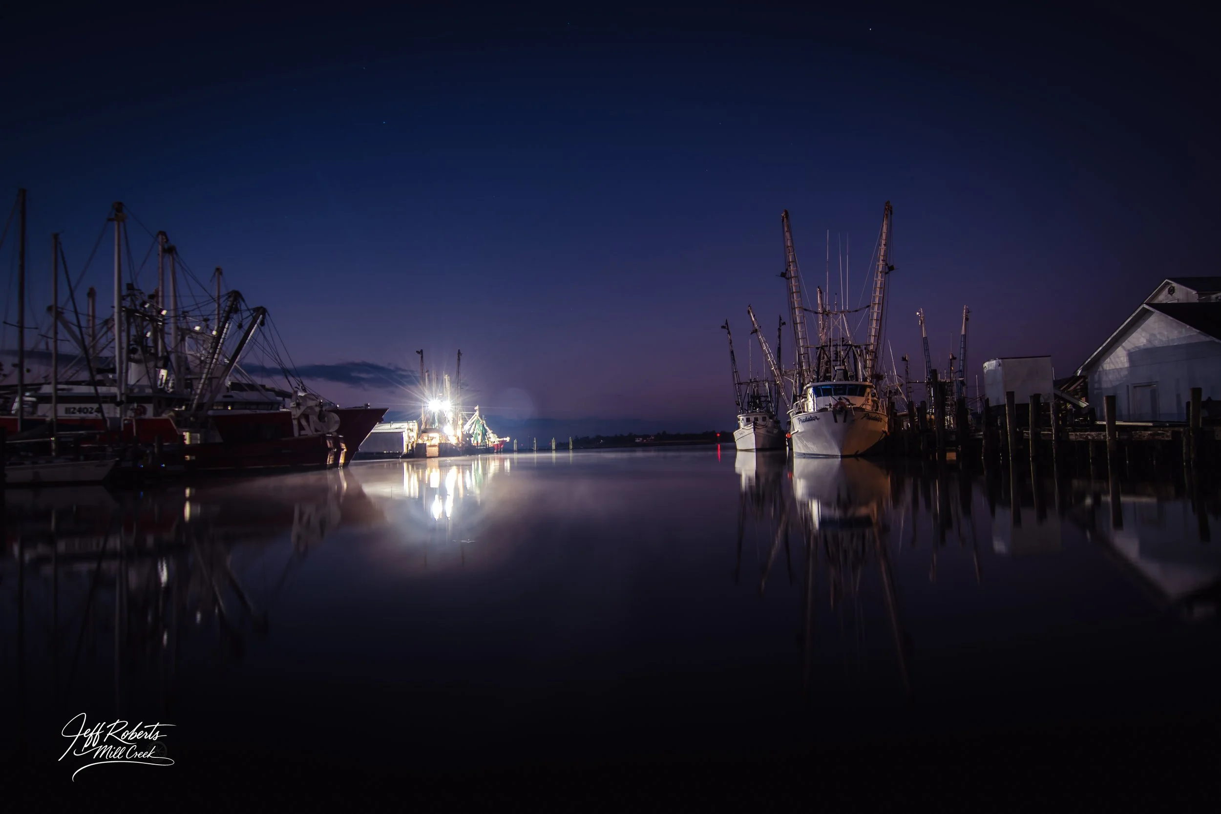 Nighttime view of boats docked at a marina with a calm water surface reflecting the boats and lights, dark sky with few stars, and a signature "Jeff Roberts Mill Creek" at the bottom left corner.