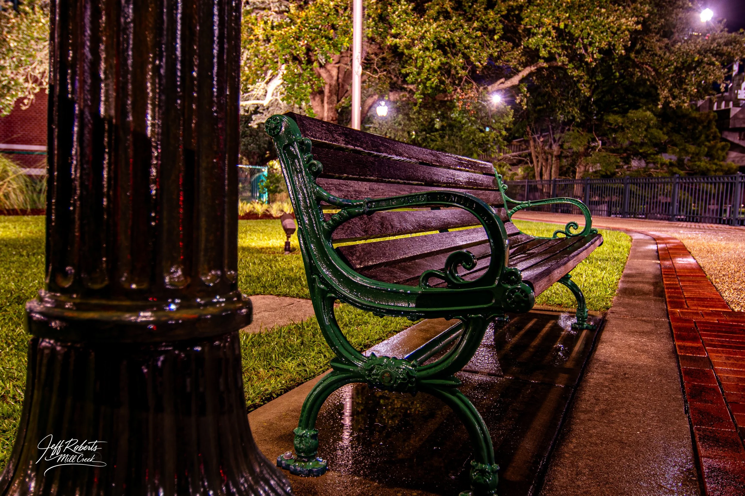 Nighttime scene of a wet park bench with metal armrests and wooden slats, illuminated by streetlights. The bench is next to a black lamppost and is situated along a curved brick and concrete pathway, with trees and a metal fence in the background.