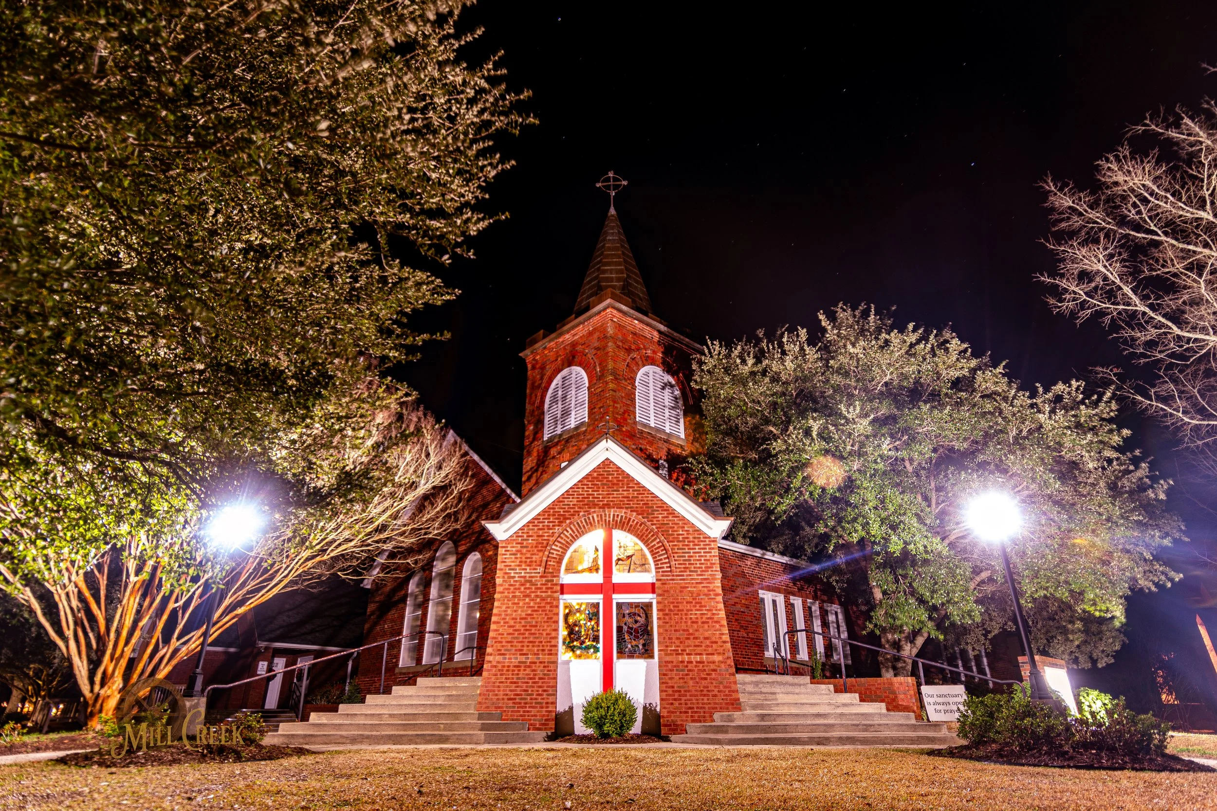 Night view of a brick church illuminated by bright outdoor lights, with stairs leading up to the entrance and trees on either side, against a dark sky.