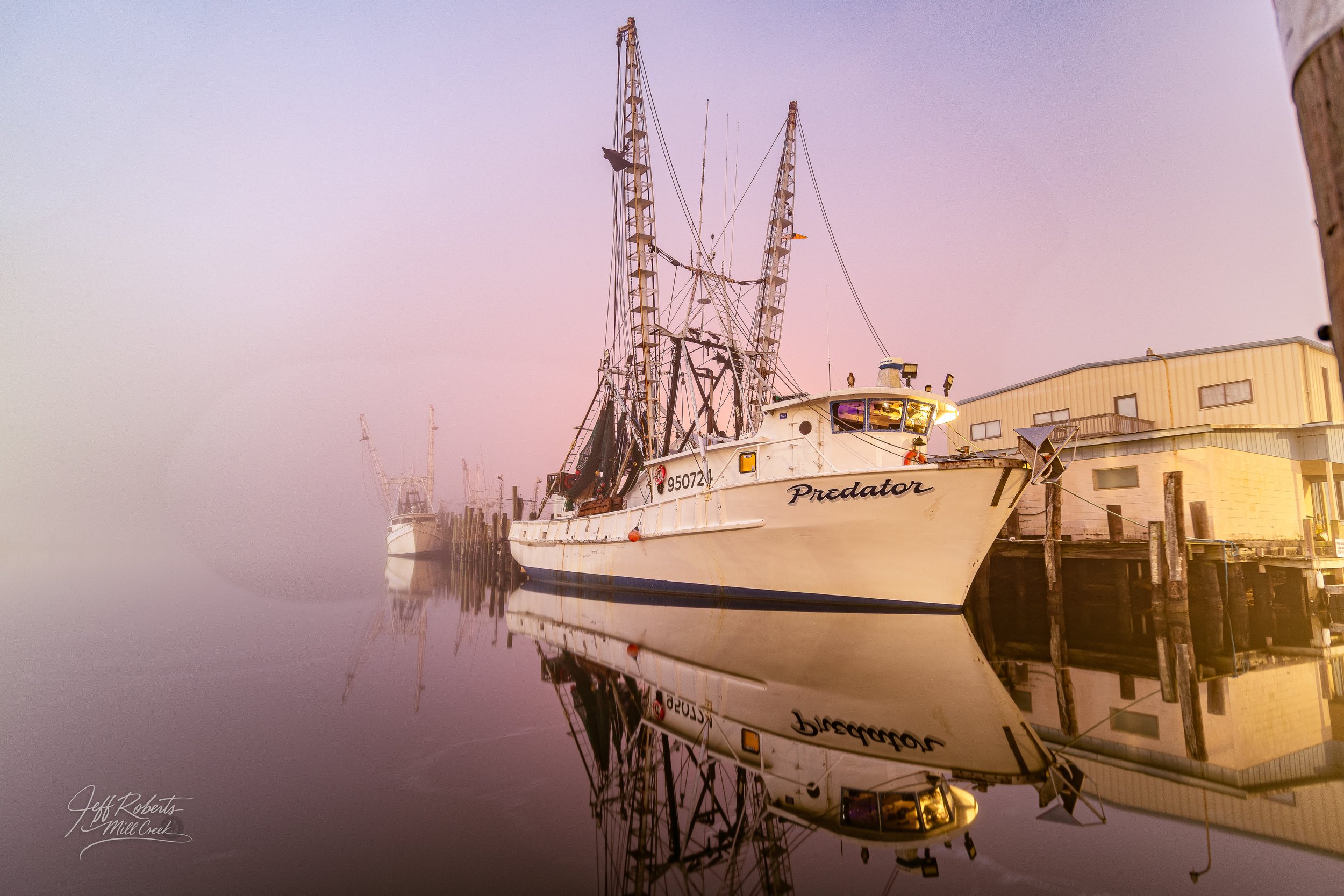 A fishing boat named Predator docked at a pier with calm water creating reflections of the boat and pier structures, during a foggy or early morning setting.