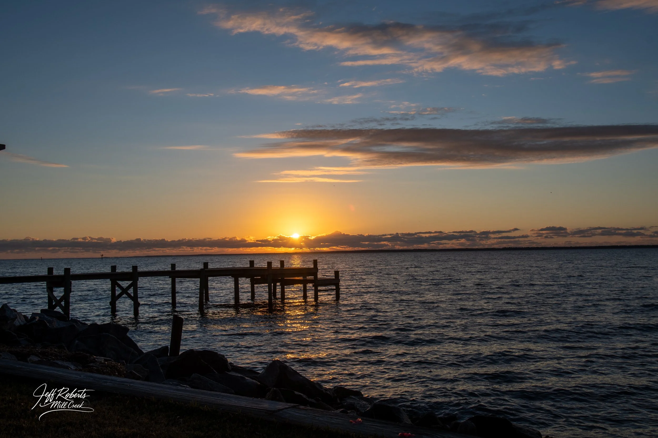 Sunset over a calm body of water with a wooden pier extending into the water and clouds in the sky.